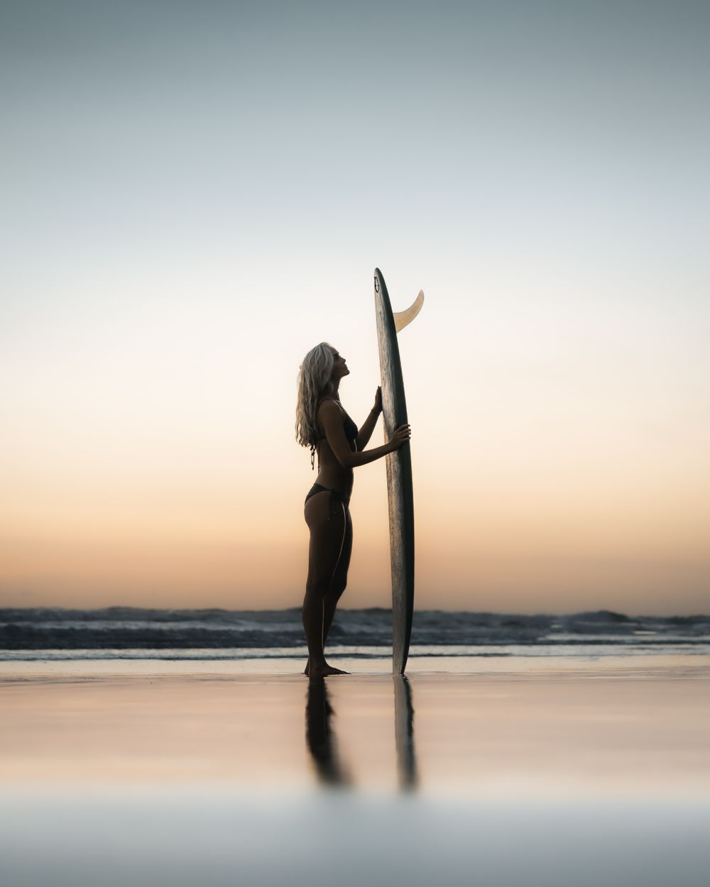 Silhouette of person holding surfboard on beach at sunset creates minimalist reflection.