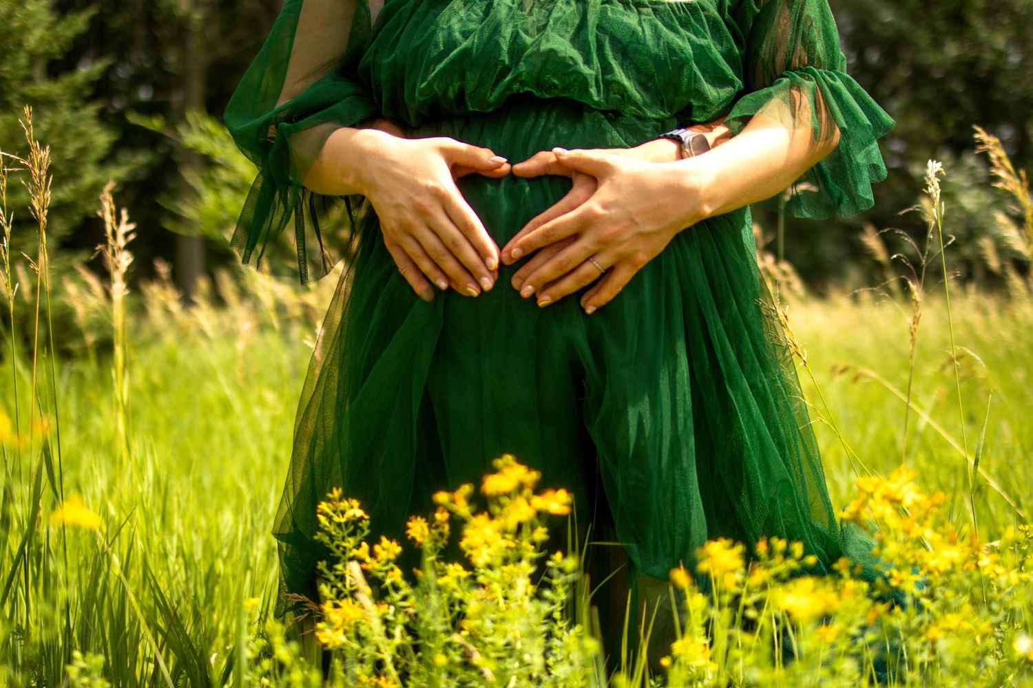 Hands rest gently on a green dress amid blooming yellow wildflowers in a sunny meadow.