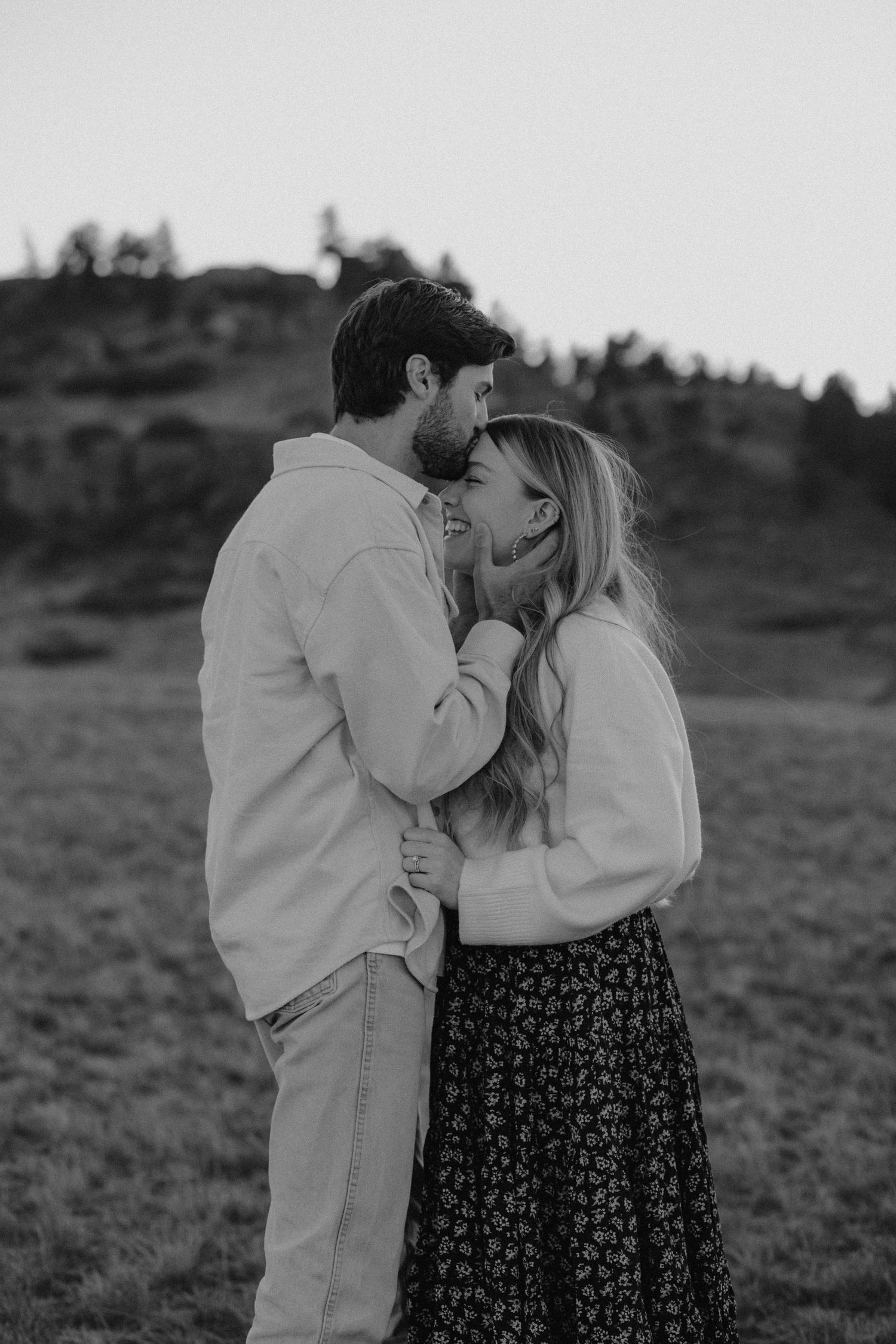 A tender black and white portrait of a romantic couple embracing outdoors in a field at sunset.