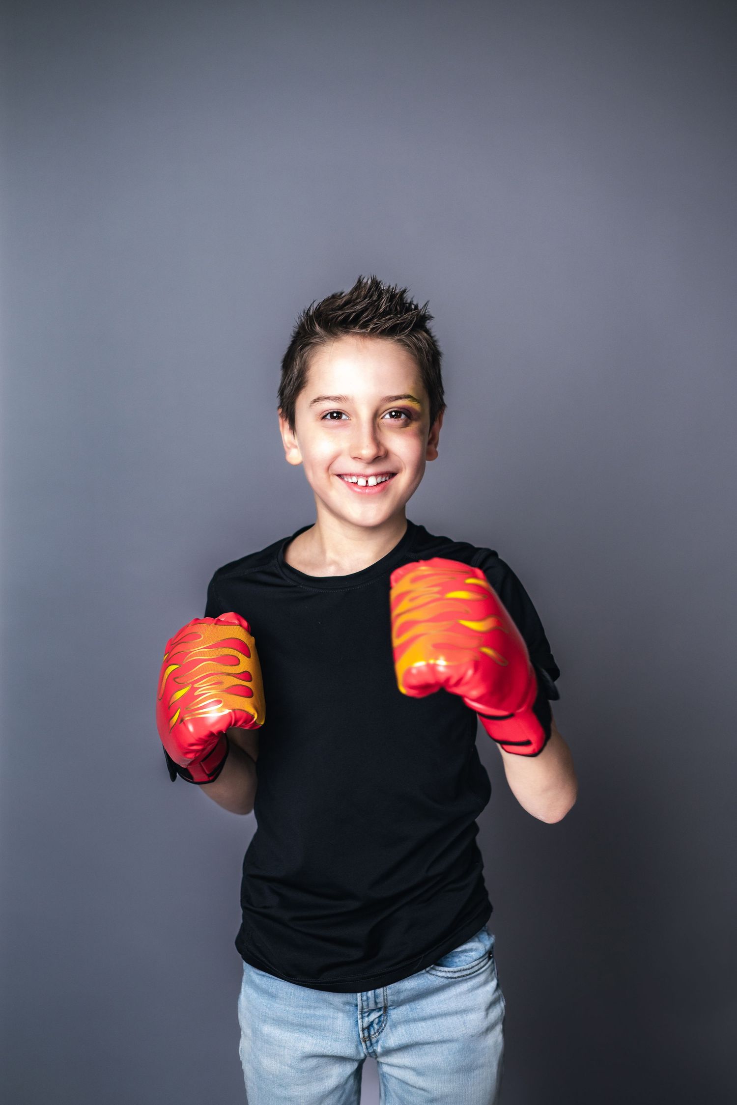 A young boxer poses with bright orange gloves against a studio backdrop wearing a black t-shirt and jeans.