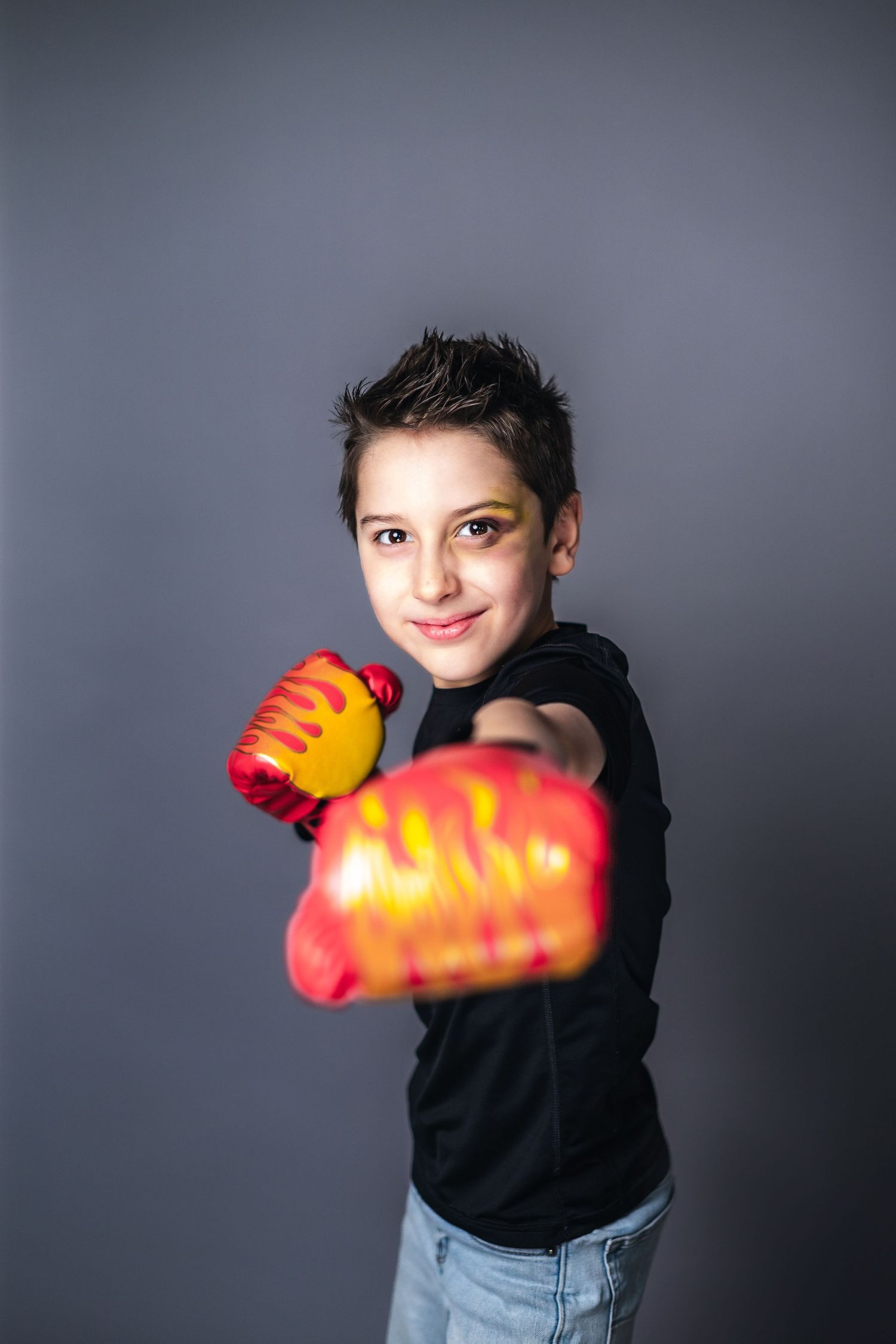 Someone wearing a black shirt and jeans strikes a playful boxing pose with bright red toy boxing gloves against a gray backdrop.
