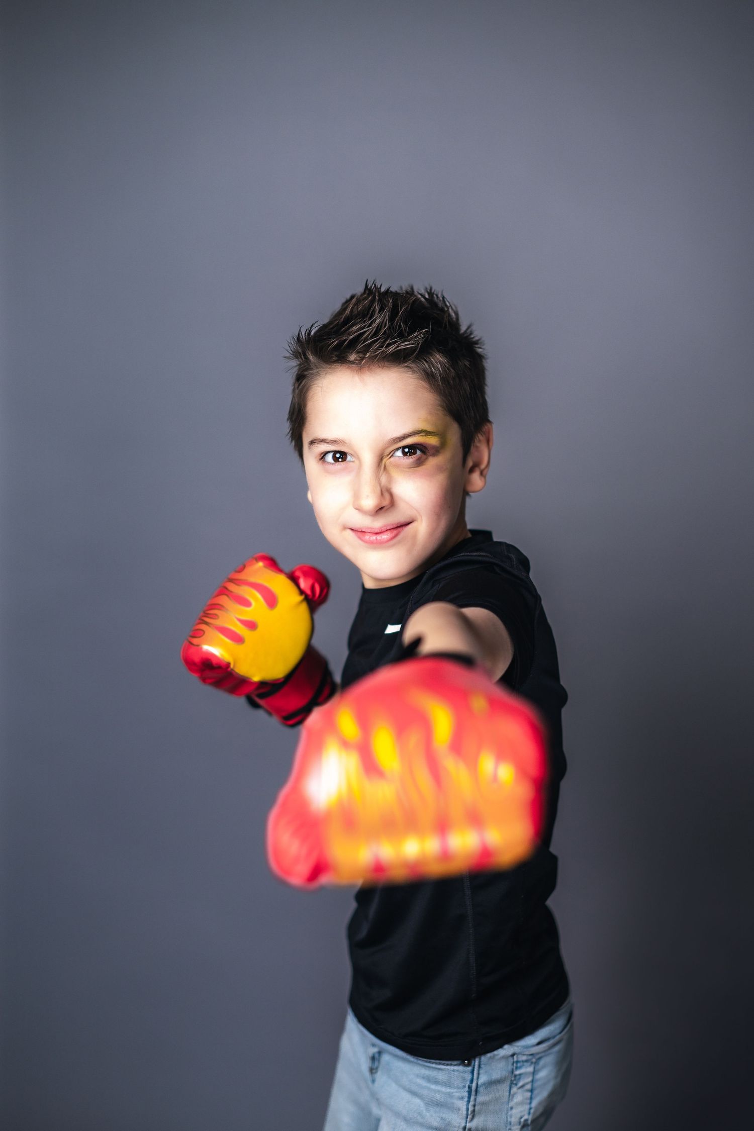 Young person wearing black hoodie and light denim jeans throwing a boxing punch with red gloves against gray background.