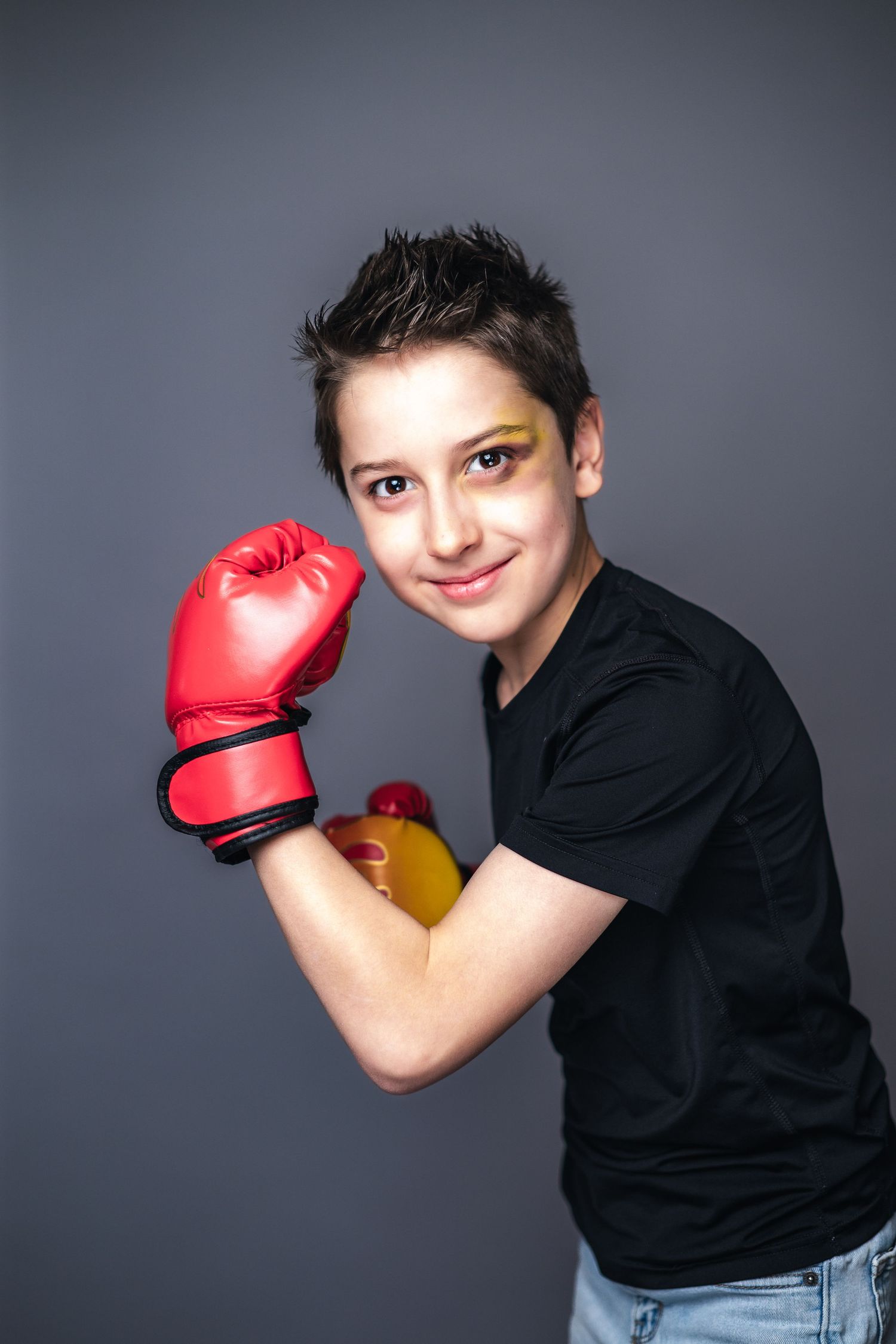 Young person in a black t-shirt wearing red boxing gloves and yellow eye makeup poses against a gray studio background.