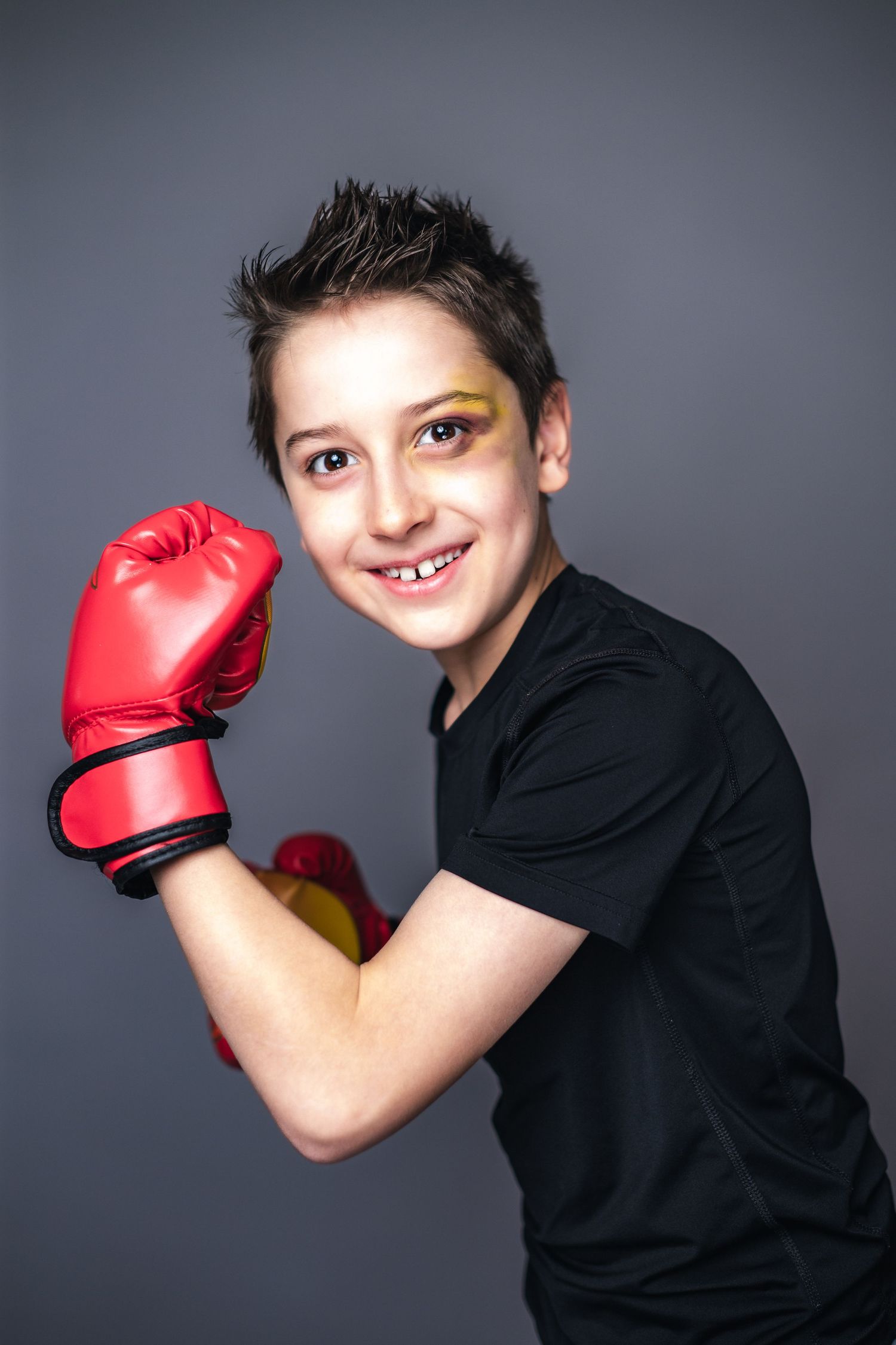 Young martial artist with spiked hair and black shirt strikes poses wearing red boxing gloves against gray background.