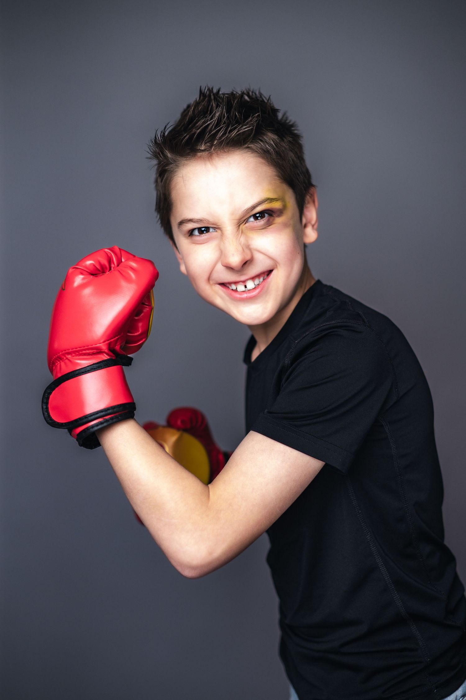 Young person in a black t-shirt smiles while wearing a red boxing glove against a gray background.