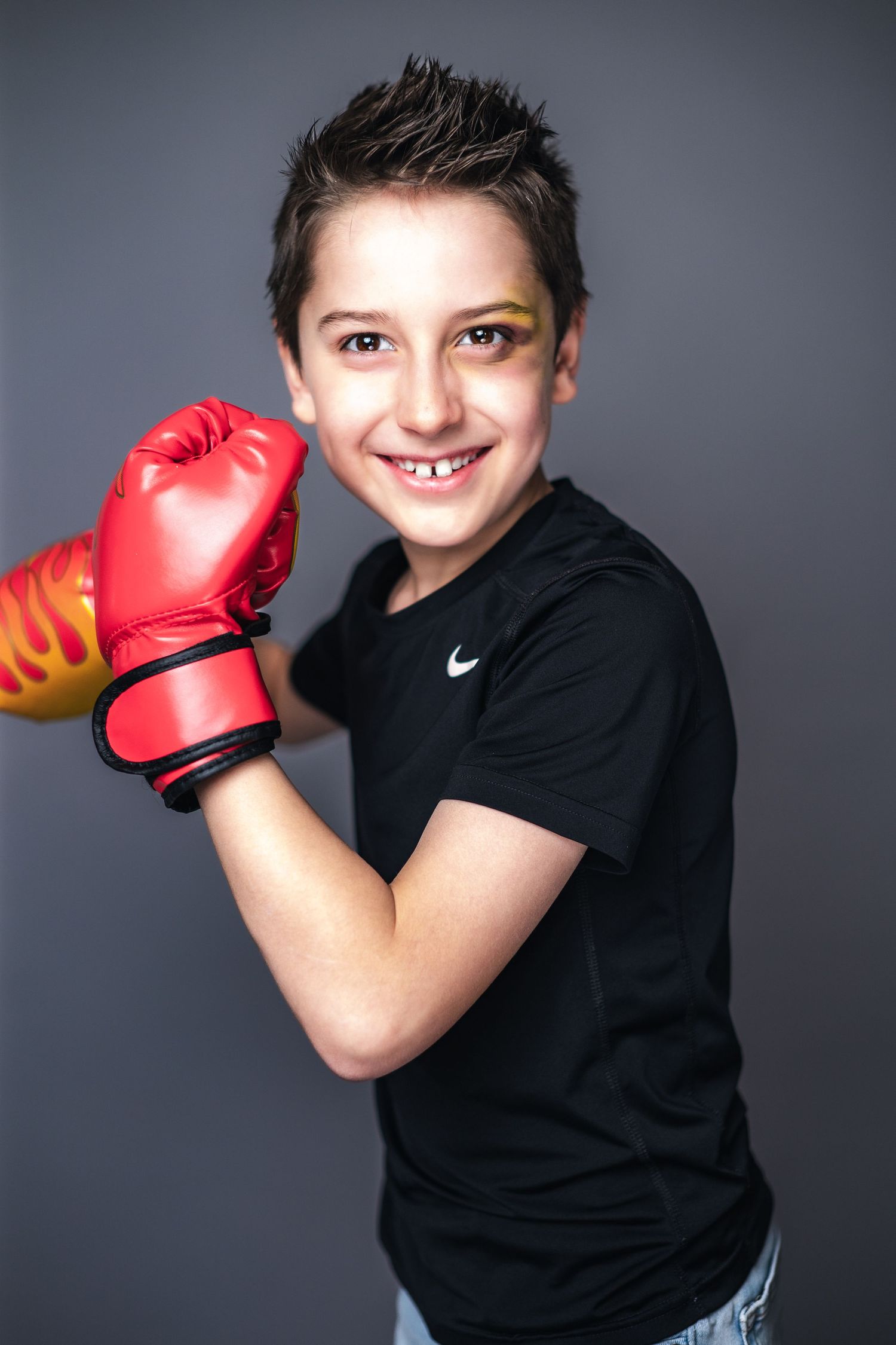 Young athlete in a black Nike t-shirt wearing red boxing gloves smiles while posing against a grey background.