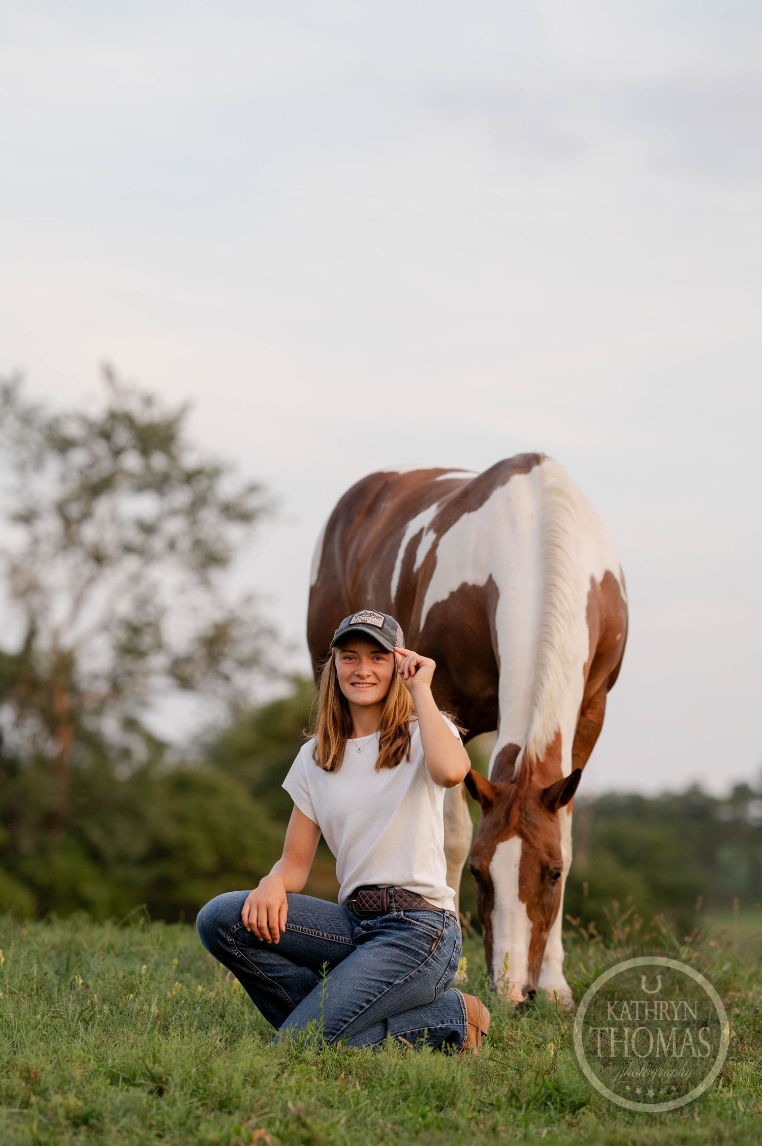 Paint horse grazes while companion in casual attire kneels nearby in grassy field.