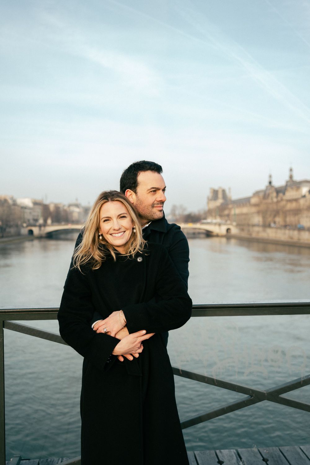 A couple embraces on a bridge overlooking the Seine River in Paris with historic buildings in the background.