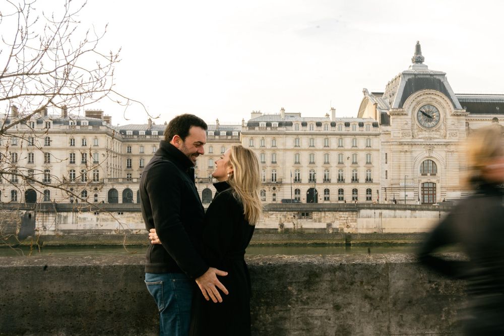 A romantic couple embraces along the Seine River in Paris with the elegant Musee d'Orsay visible in the background.