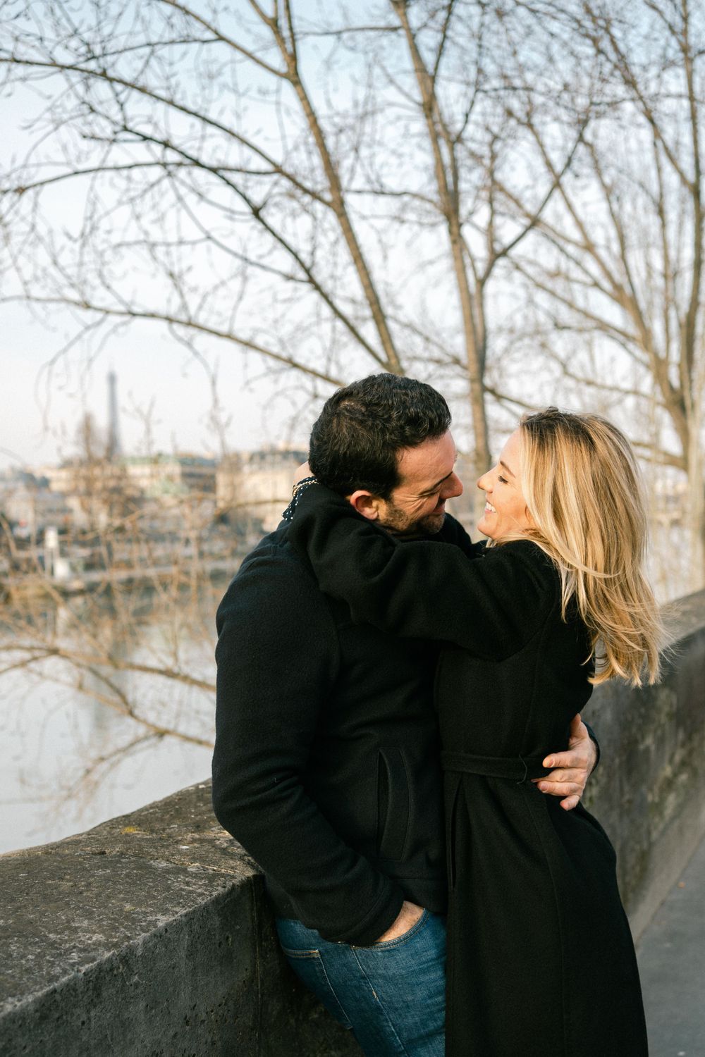 A romantic couple in winter coats share an intimate moment along a stone wall with bare trees and city views in background.