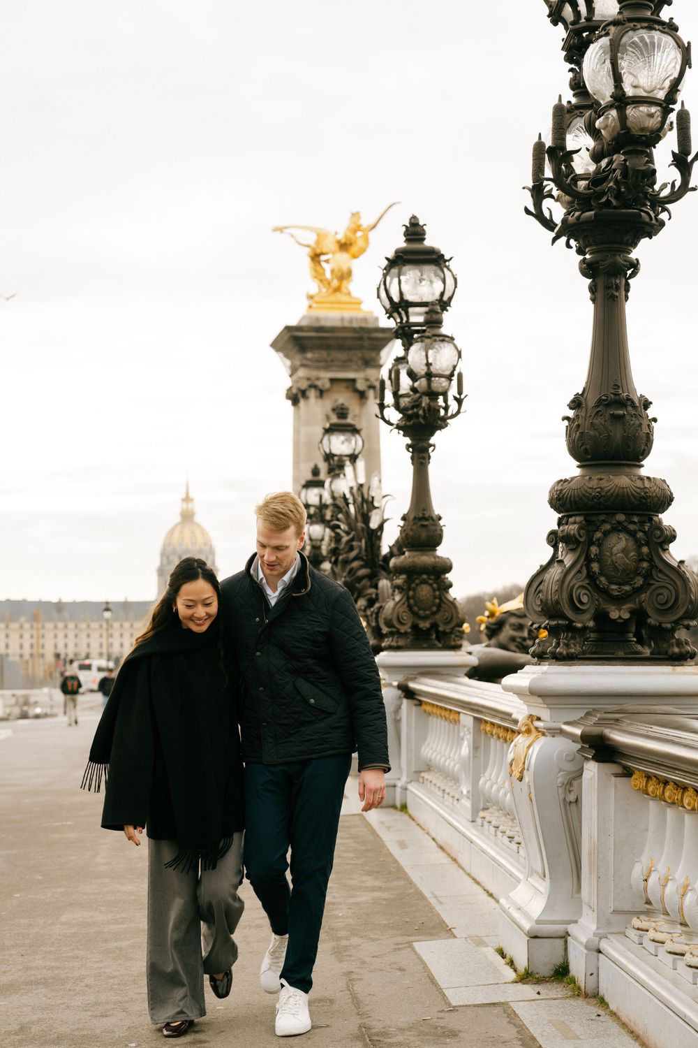 A couple strolls along the Pont Alexandre III bridge in Paris with ornate lampposts and golden statues in the background.
