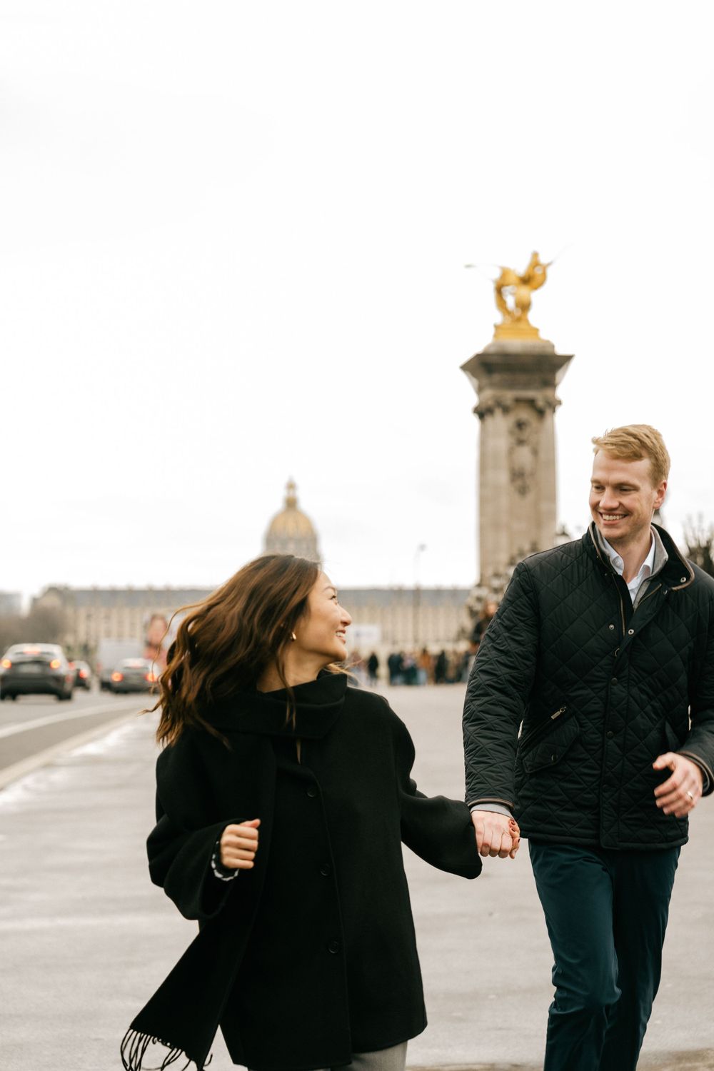 Couple walking and smiling together on a bridge in Paris with ornate architecture in the background.