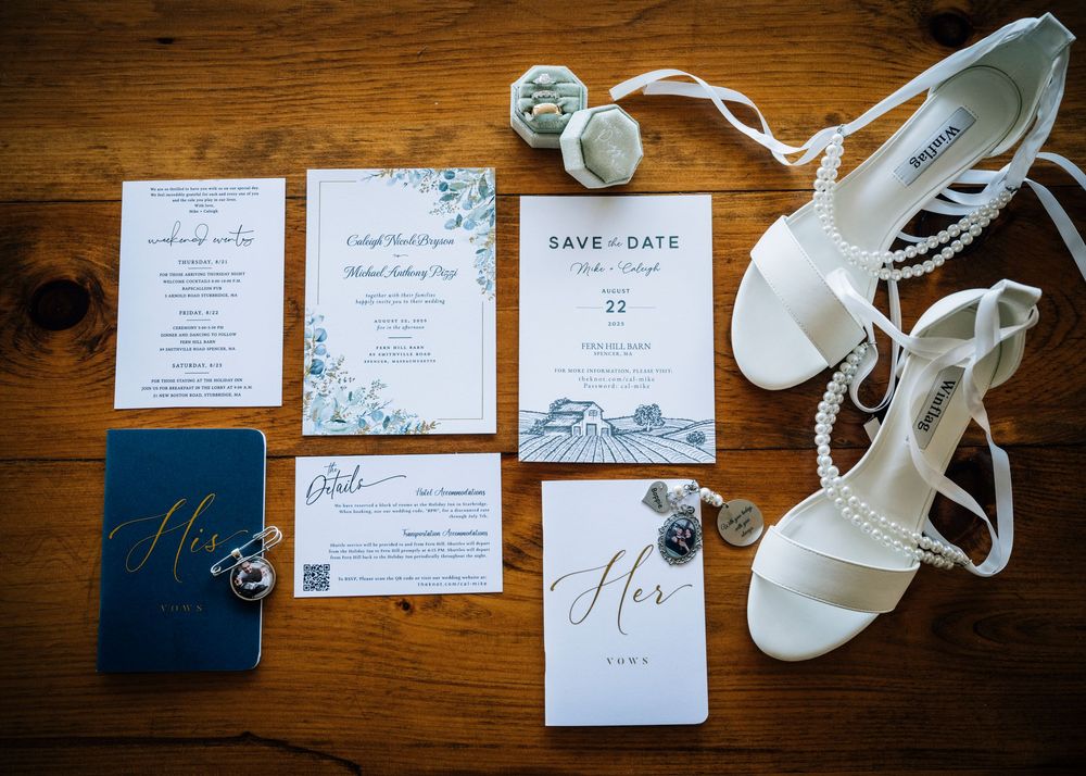 Wedding details flatlay featuring invitations, rings, and white bridal shoes on wooden surface.