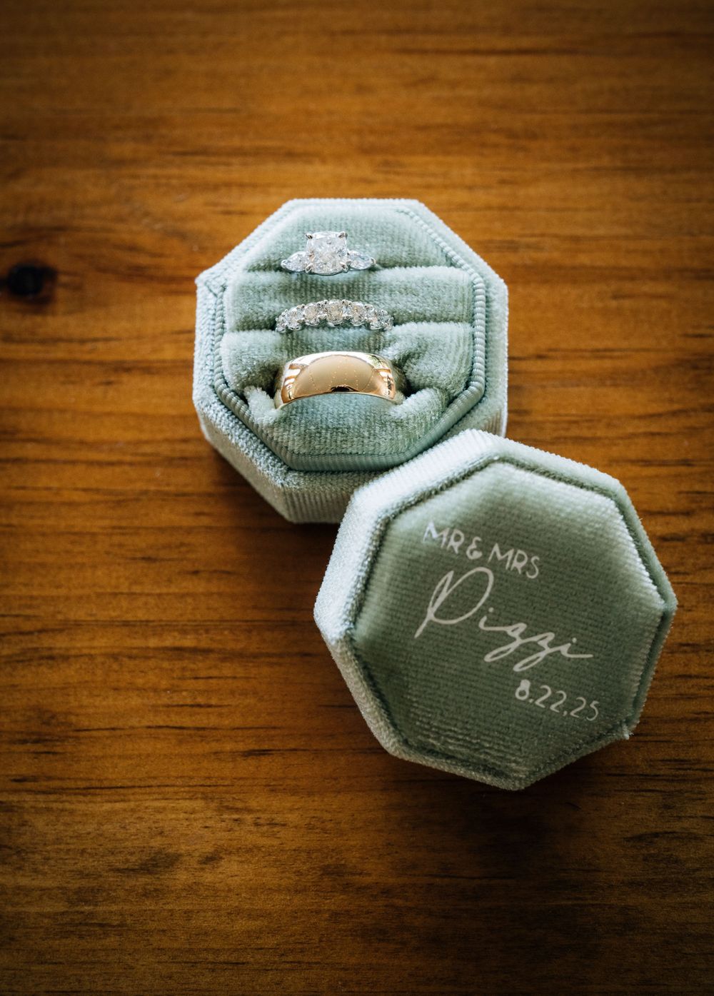 Two mint green velvet octagonal ring boxes with wedding details displayed on wooden surface.