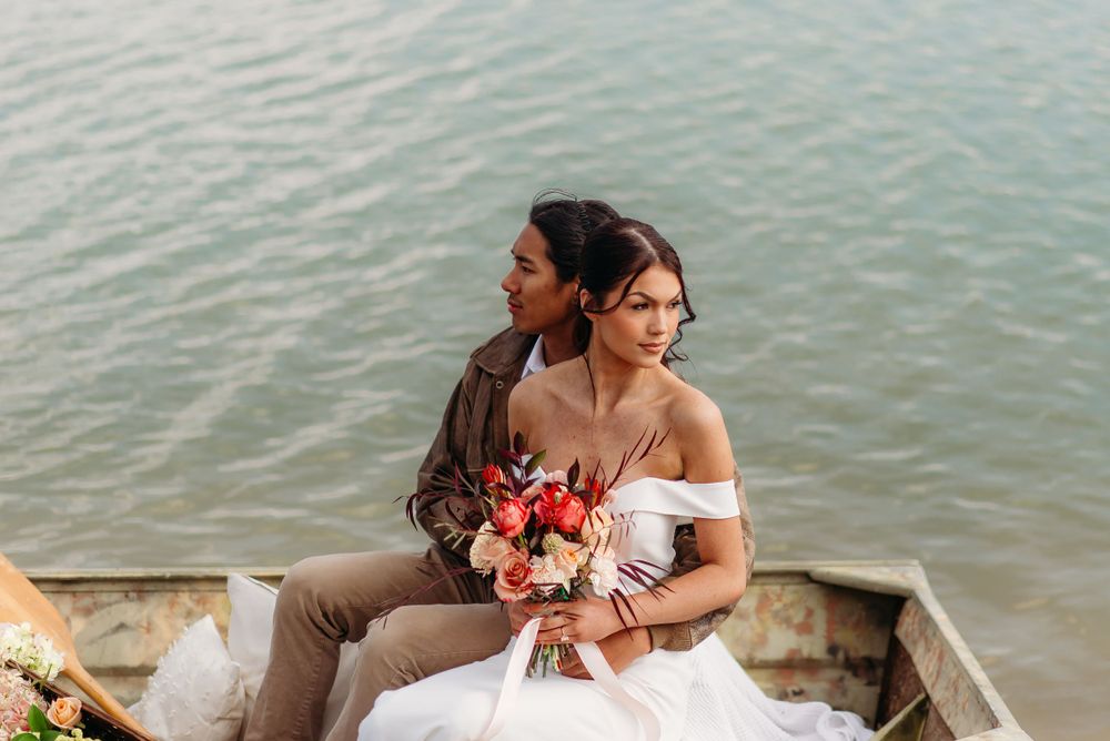A romantic couple in wedding attire sitting by the water holding a coral and pink bouquet.