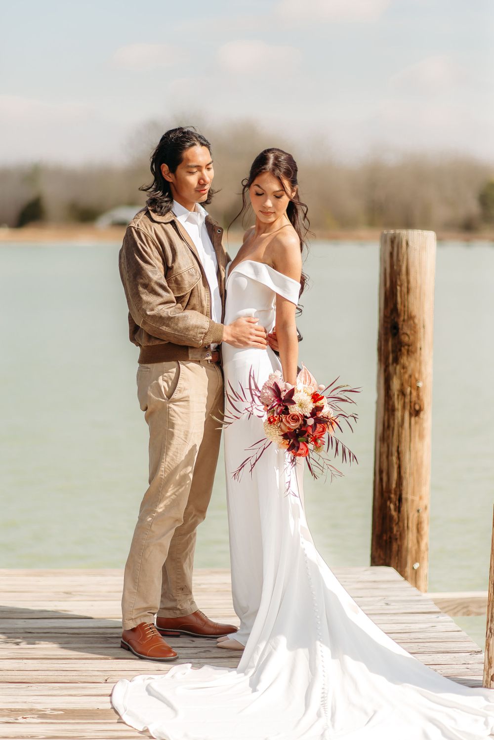 Elegant couple poses on wooden dock, bride in white dress holding coral bouquet.