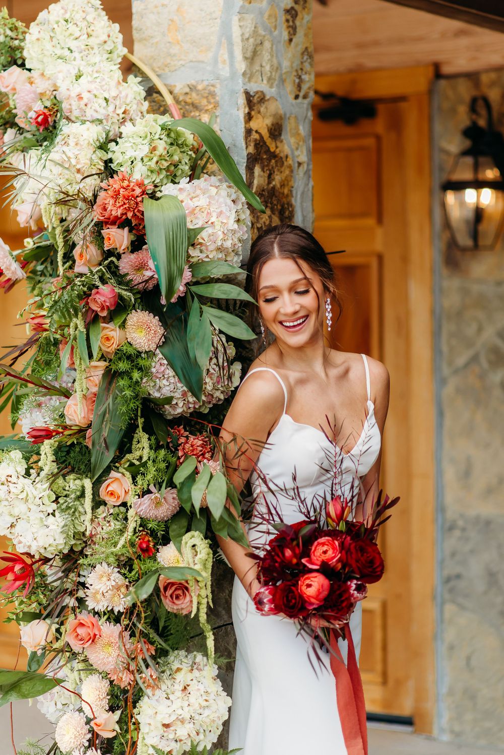 A person in a white wedding dress smiles while holding a red bouquet next to lush floral arrangements.