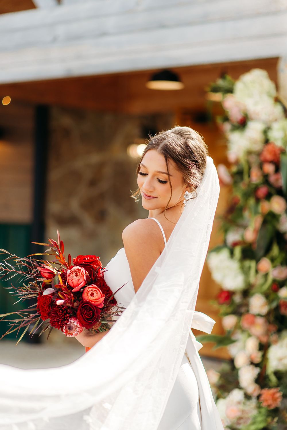 A flowing white wedding dress and veil catch the breeze as a person holds a red bouquet.