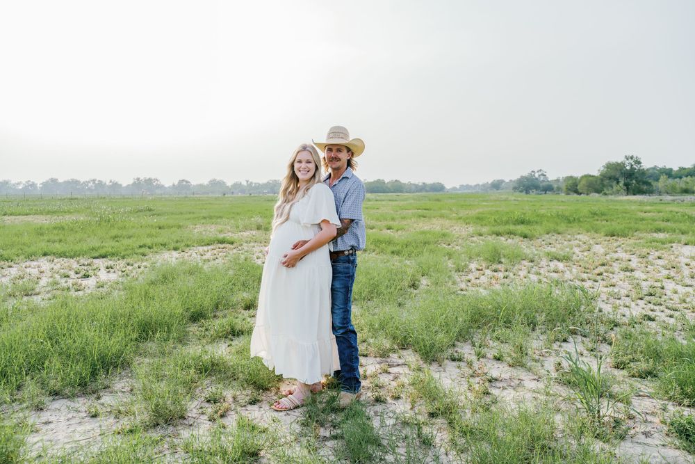 Pregnant couple posing in a green field, woman in white dress, man in cowboy hat and jeans, sunny outdoor maternity photo.