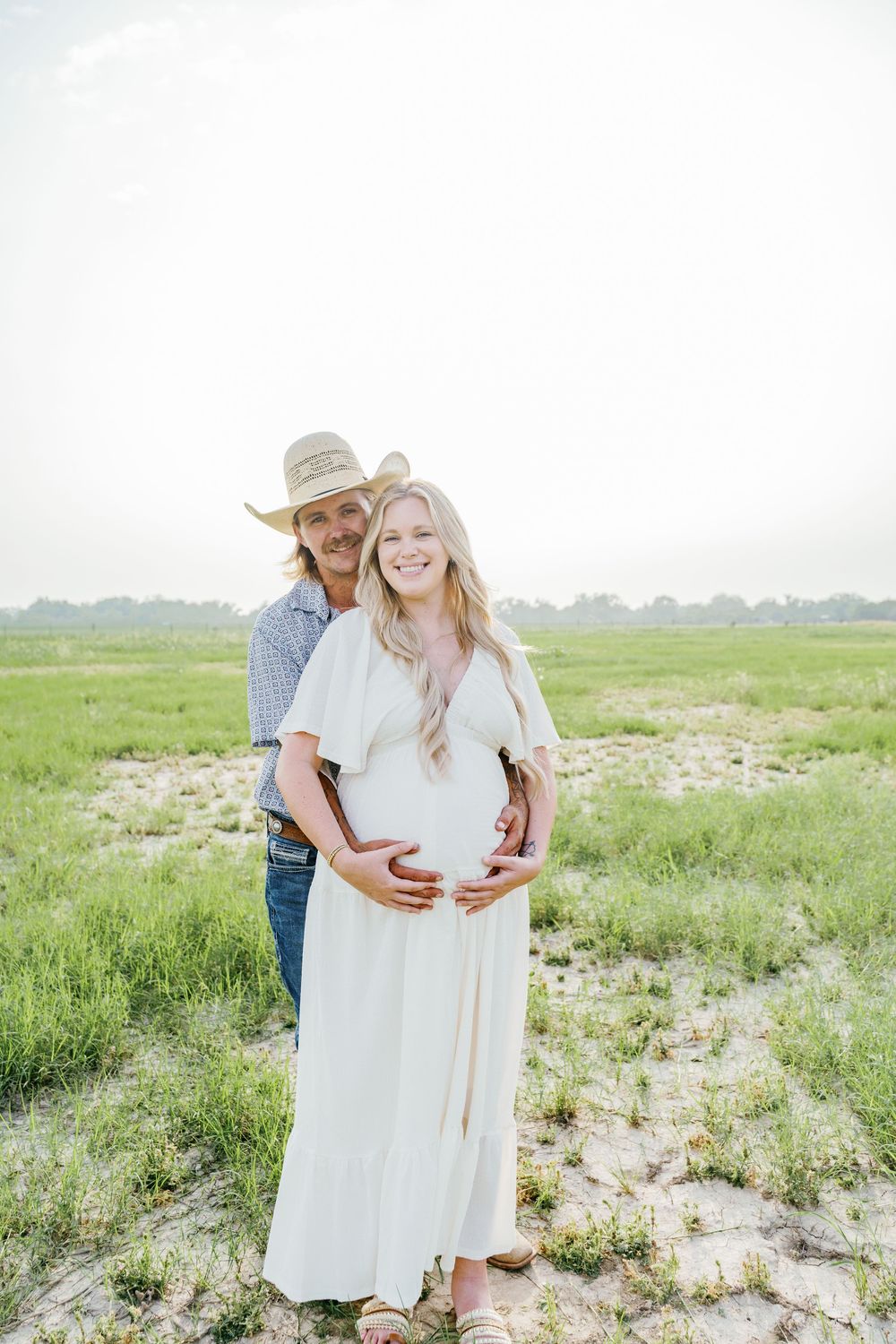 Couple posing in green field, man in cowboy hat embracing pregnant woman in white dress outdoors.