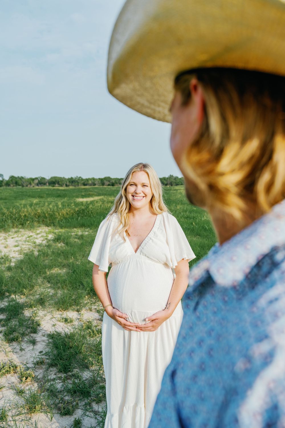 Smiling pregnant woman in white maxi dress cradling baby bump in sunny green field.