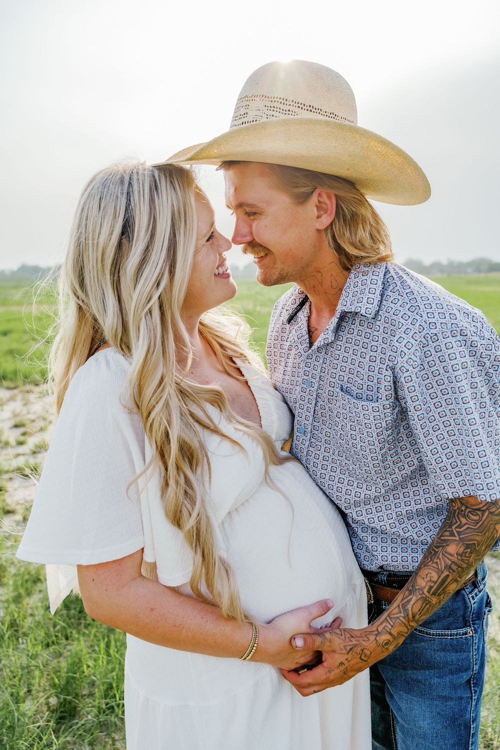 Expecting couple in white dress and cowboy hat sharing tender moment in field.