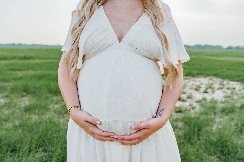 Pregnant woman in white dress cradling baby bump outdoors in green field during maternity photo session.