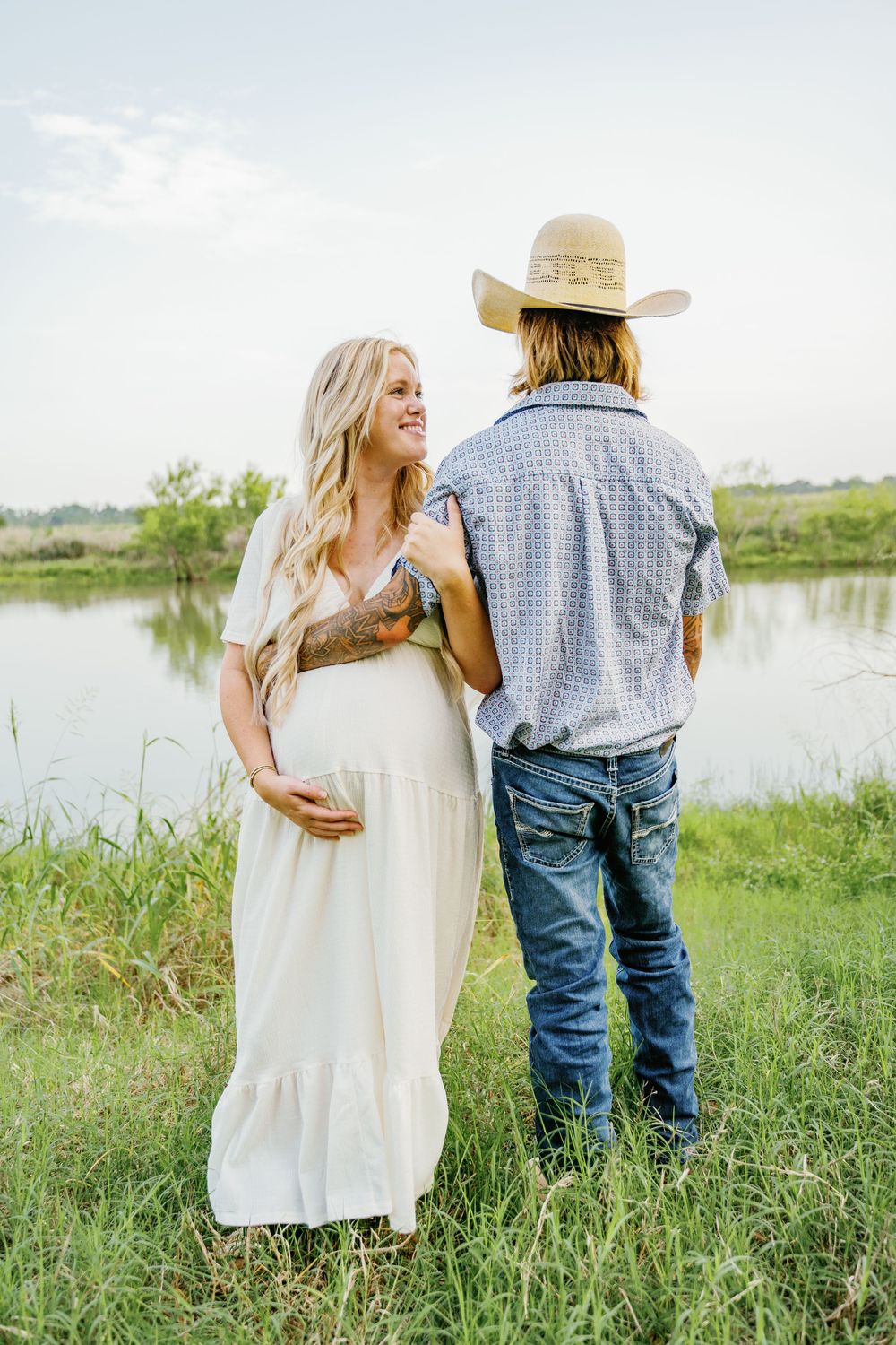 Pregnant woman in white dress smiles at tattooed partner in cowboy hat by pond.