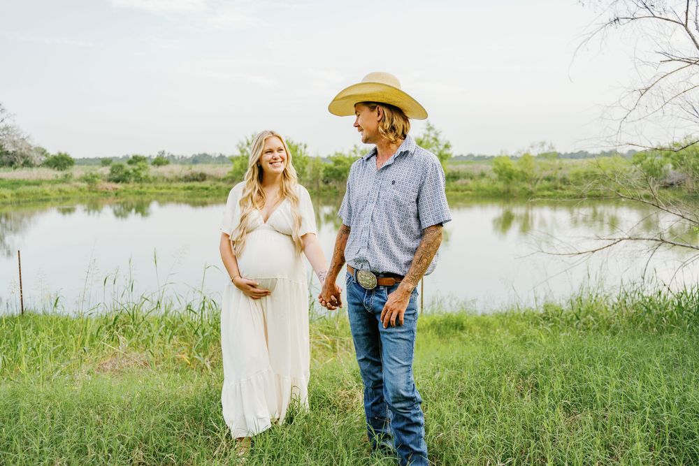 Pregnant woman in white dress holds hands with cowboy hat-wearing man by a serene pond in a maternity photo.