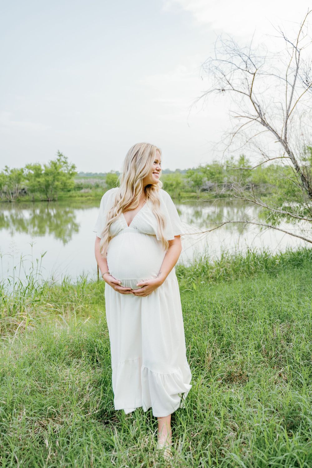 Pregnant woman in white dress cradling baby bump near a serene pond surrounded by lush green grass.