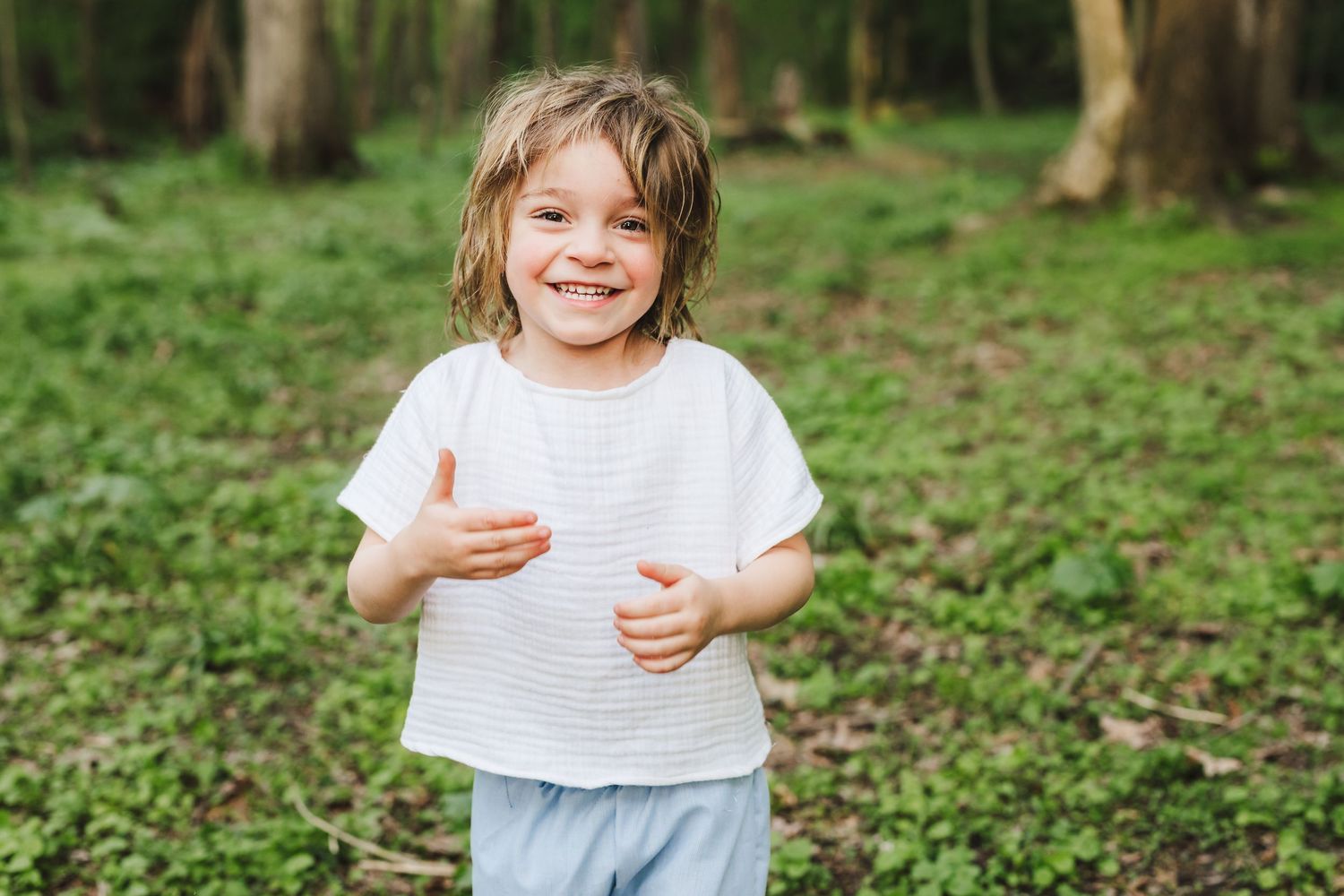 A joyful child in a white shirt stands in a green forest setting, smiling brightly.