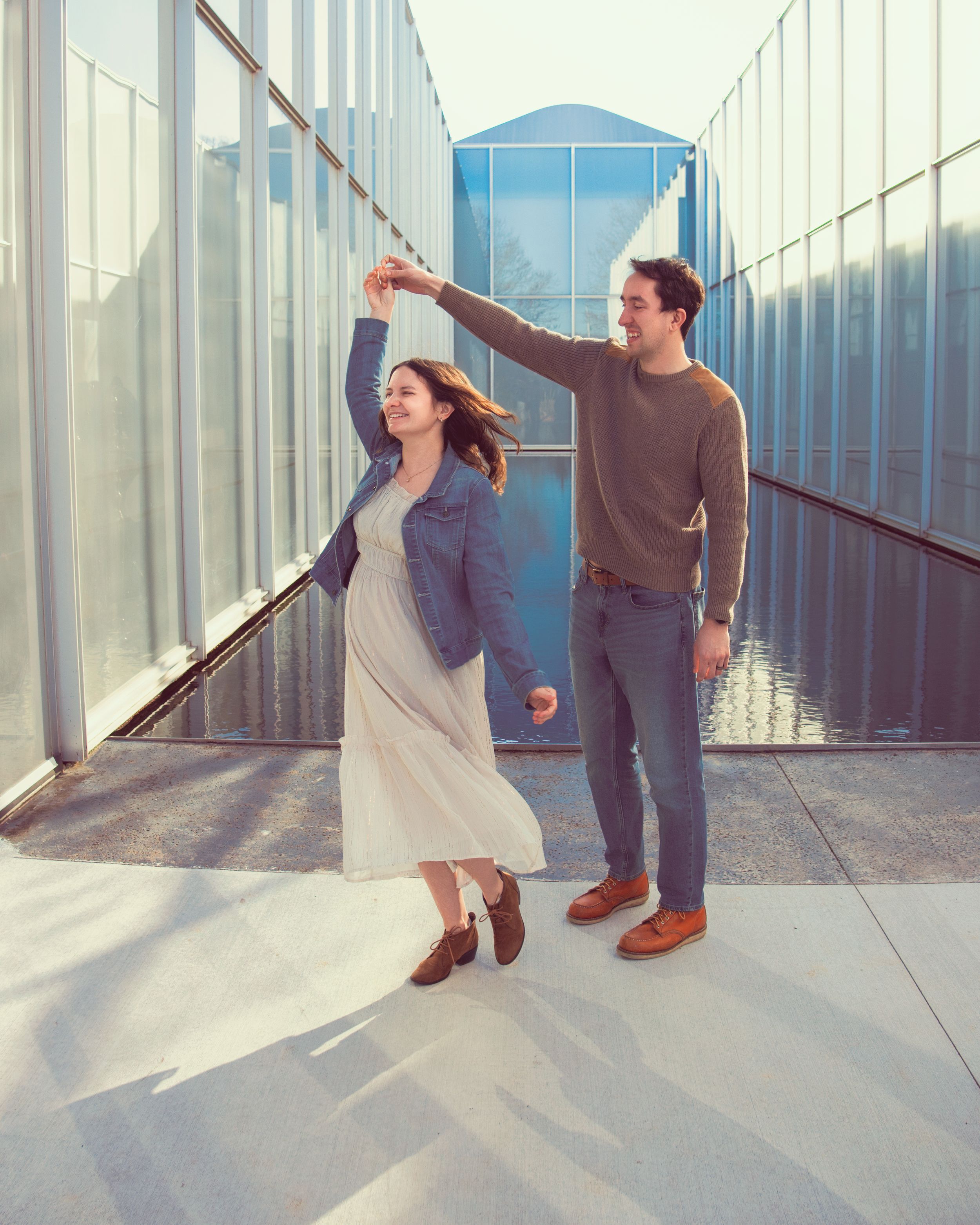 A couple dancing in a sunlit corridor between glass walls with a blue sky visible at the end.