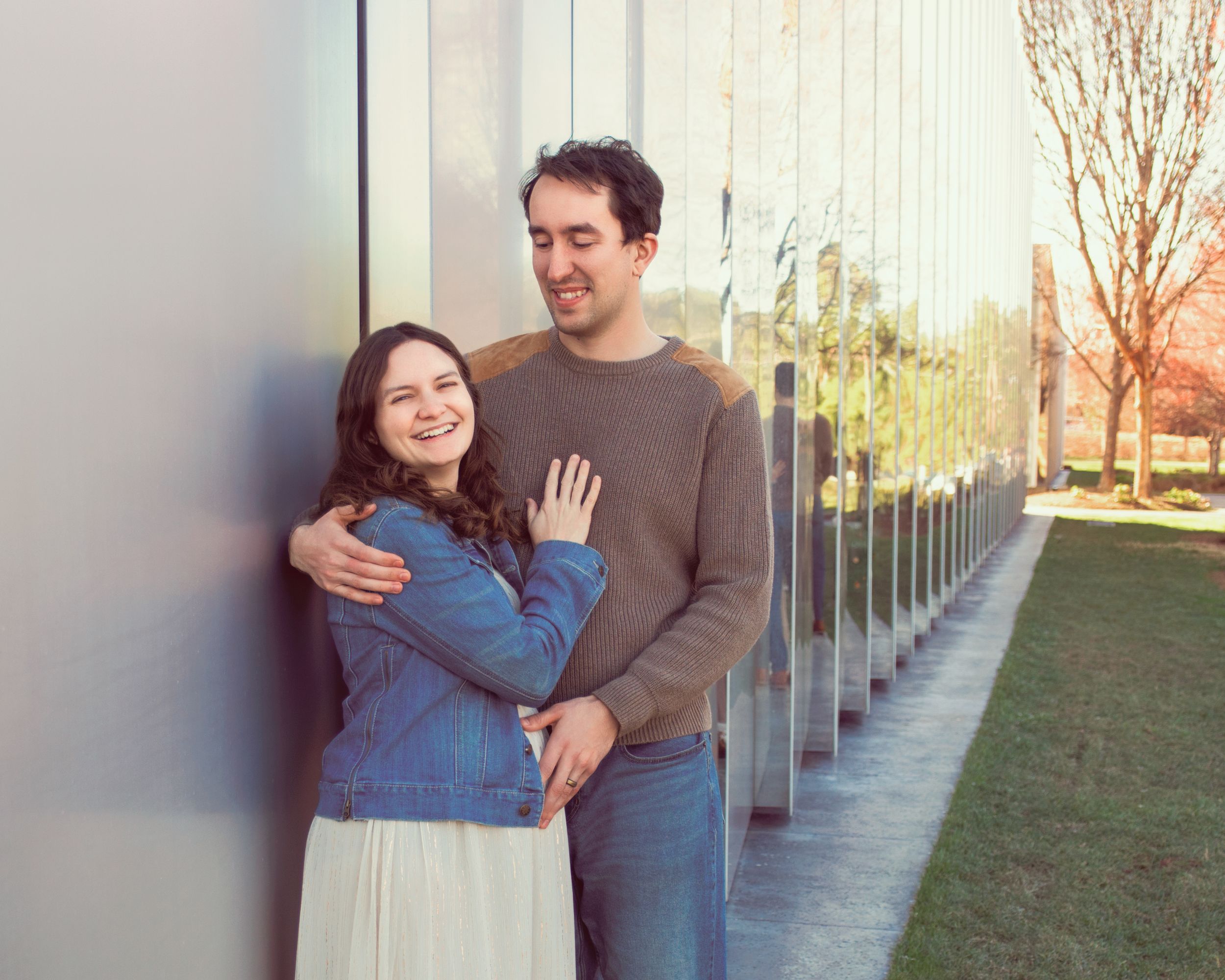 A couple stands embracing against a wall outdoors with glass panels and trees visible in the background.
