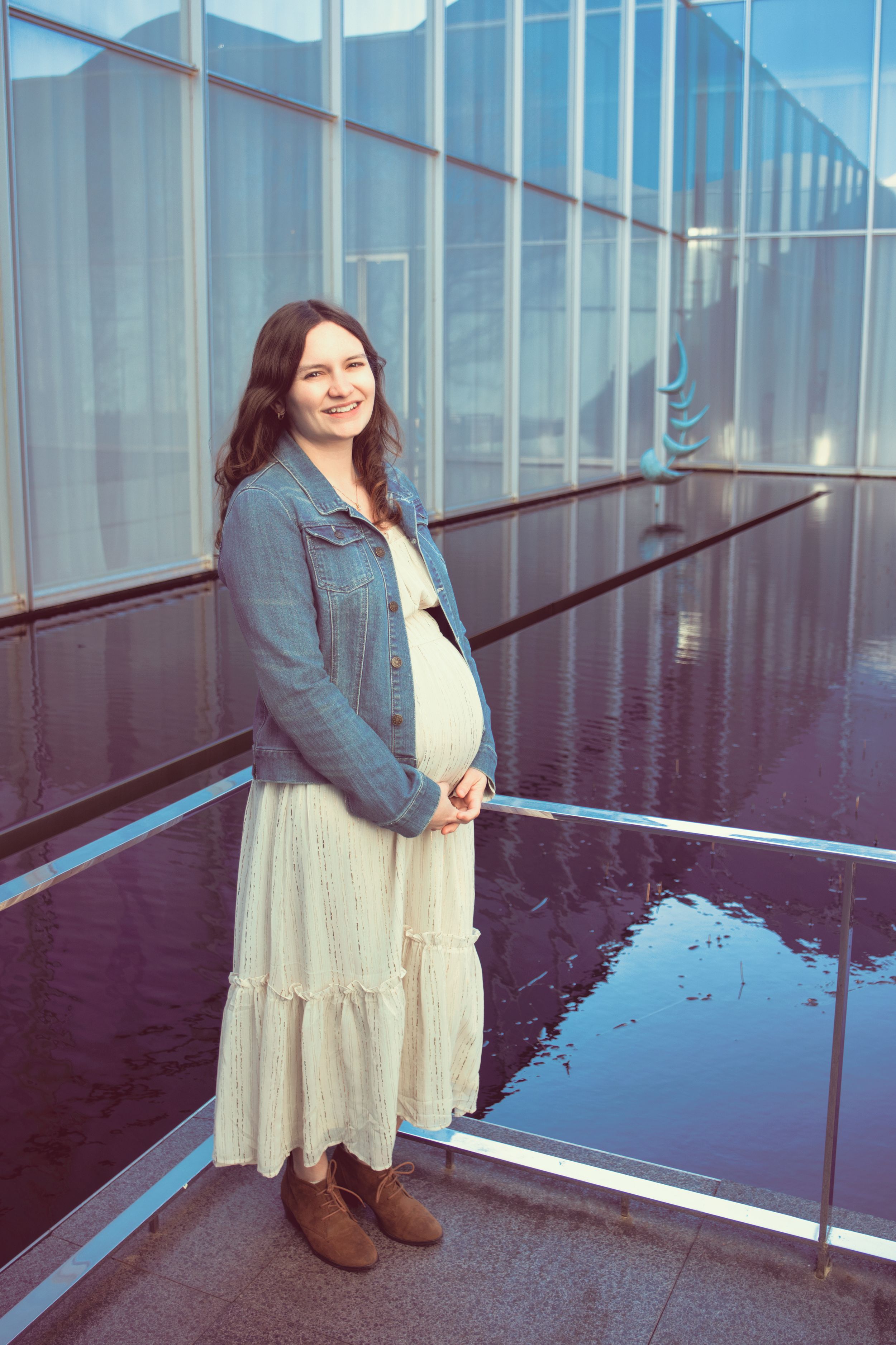An expectant parent in a denim jacket and white tiered dress stands at a glass building interior with reflective floors.
