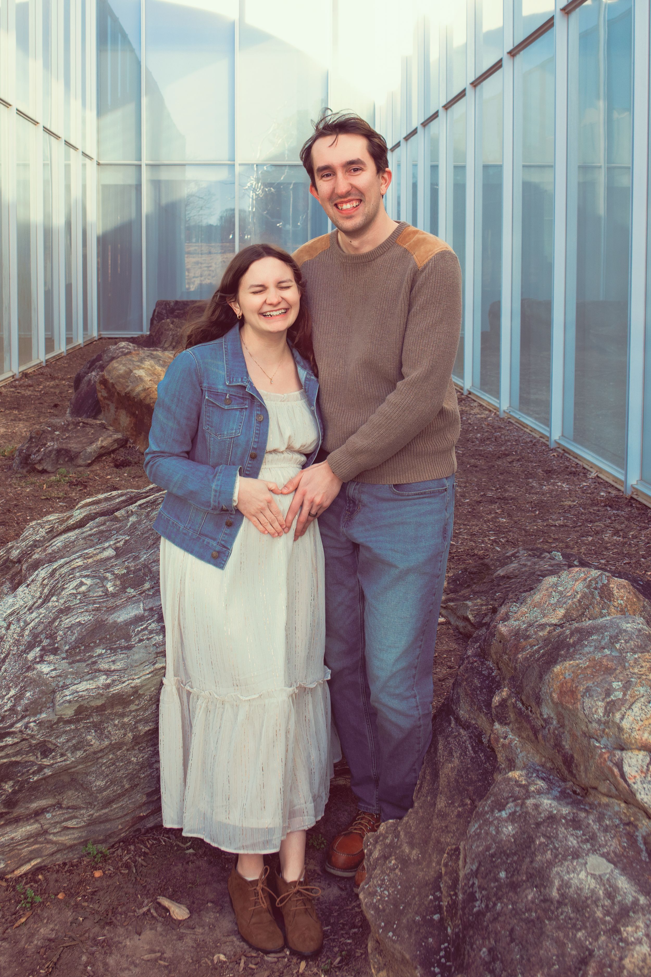 A couple stands together on rocky terrain outside a glass-walled building on an overcast day.