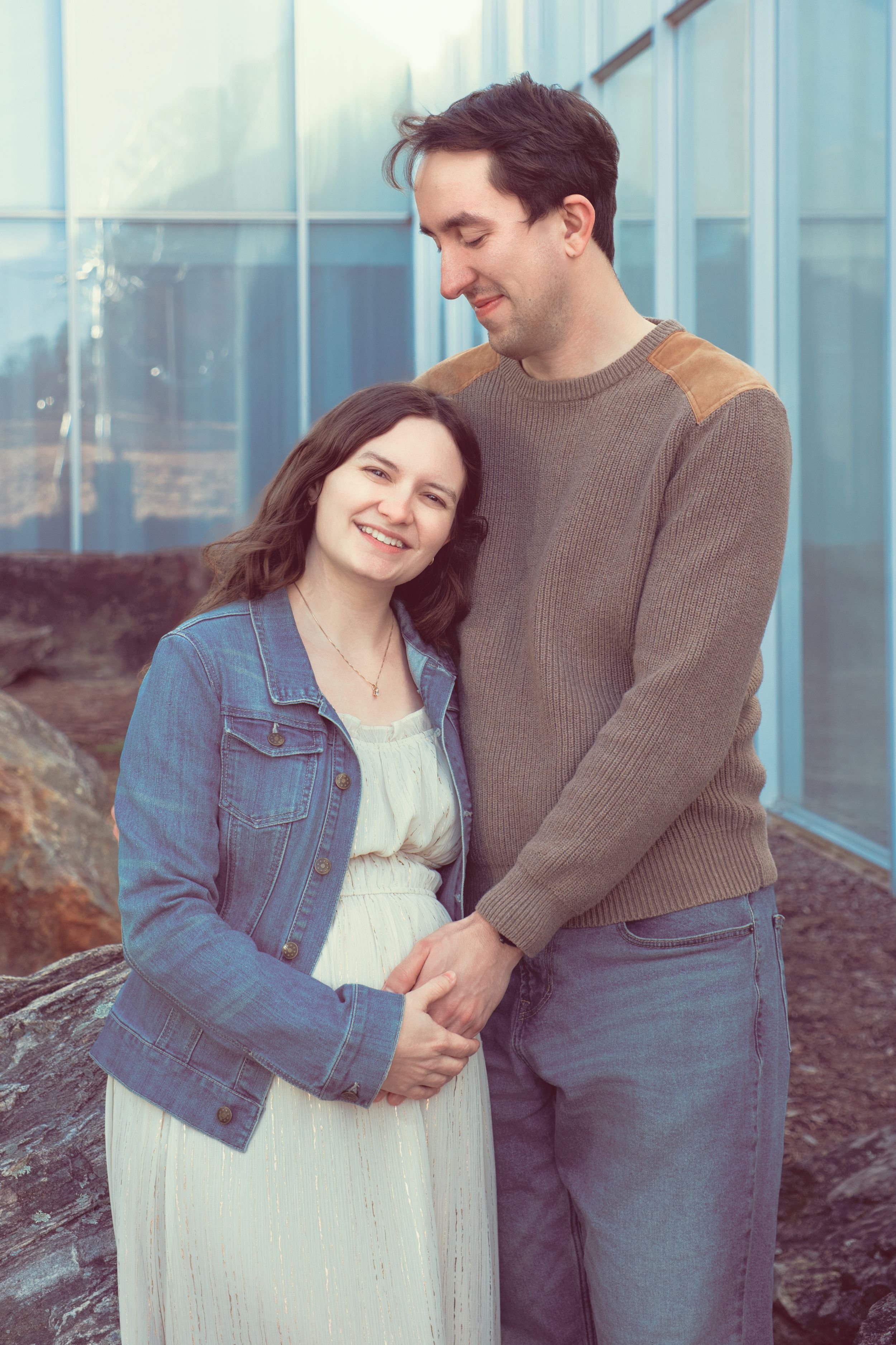 A couple stands together outdoors, with a pregnant woman in a white dress and denim jacket holding her baby bump.