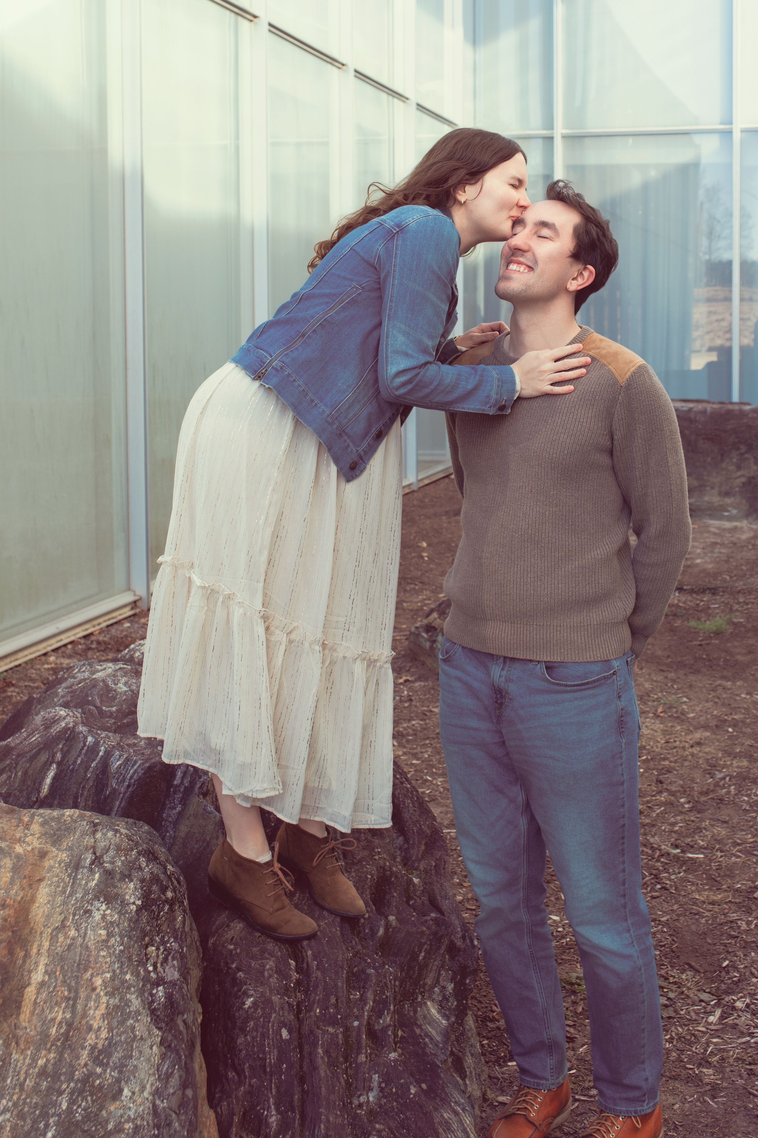 A couple shares a tender moment outdoors, with one standing on a rock to reach the other for a sweet gesture.