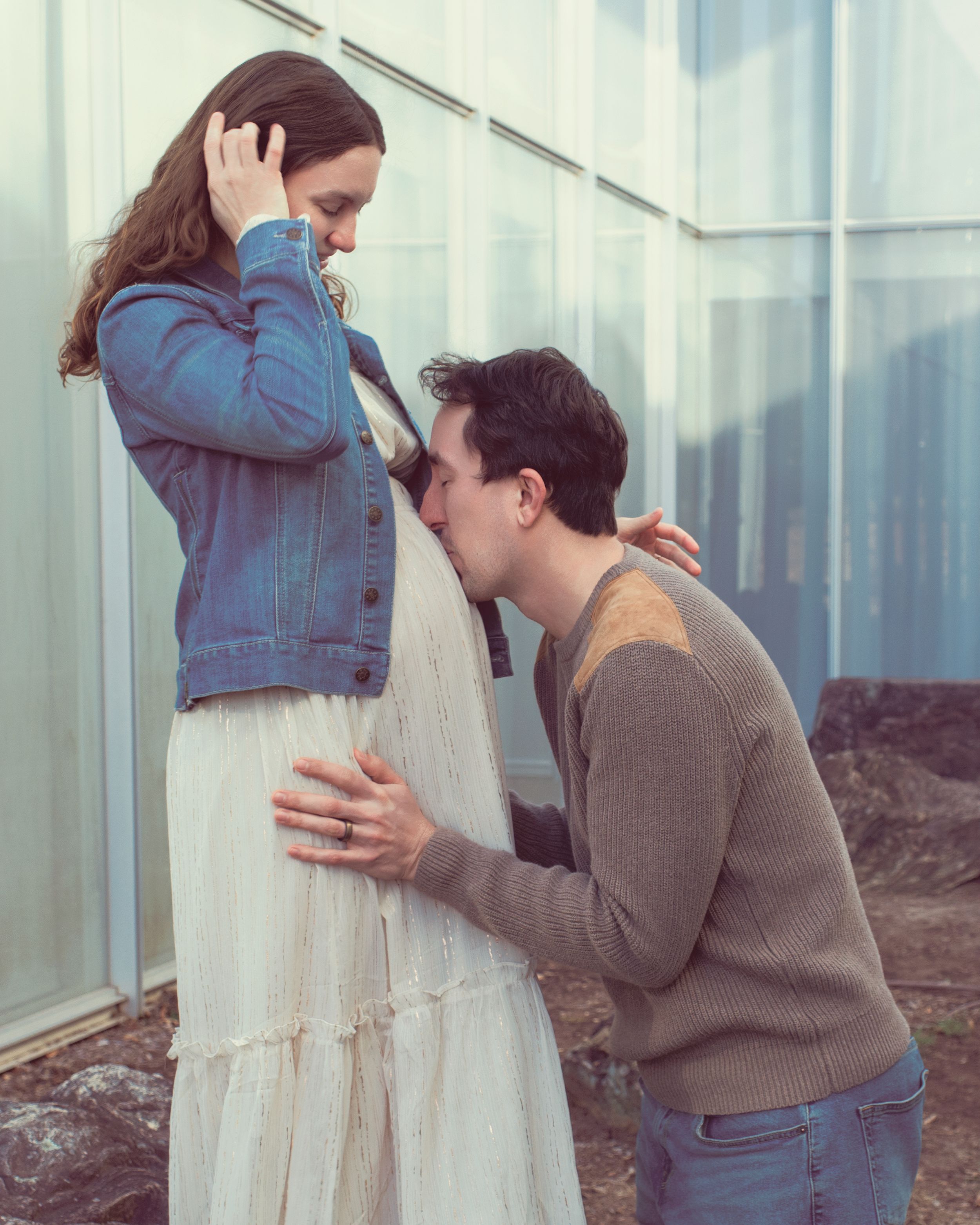 A sweet candid moment shared between a couple near a window, with her in a denim jacket and white dress.