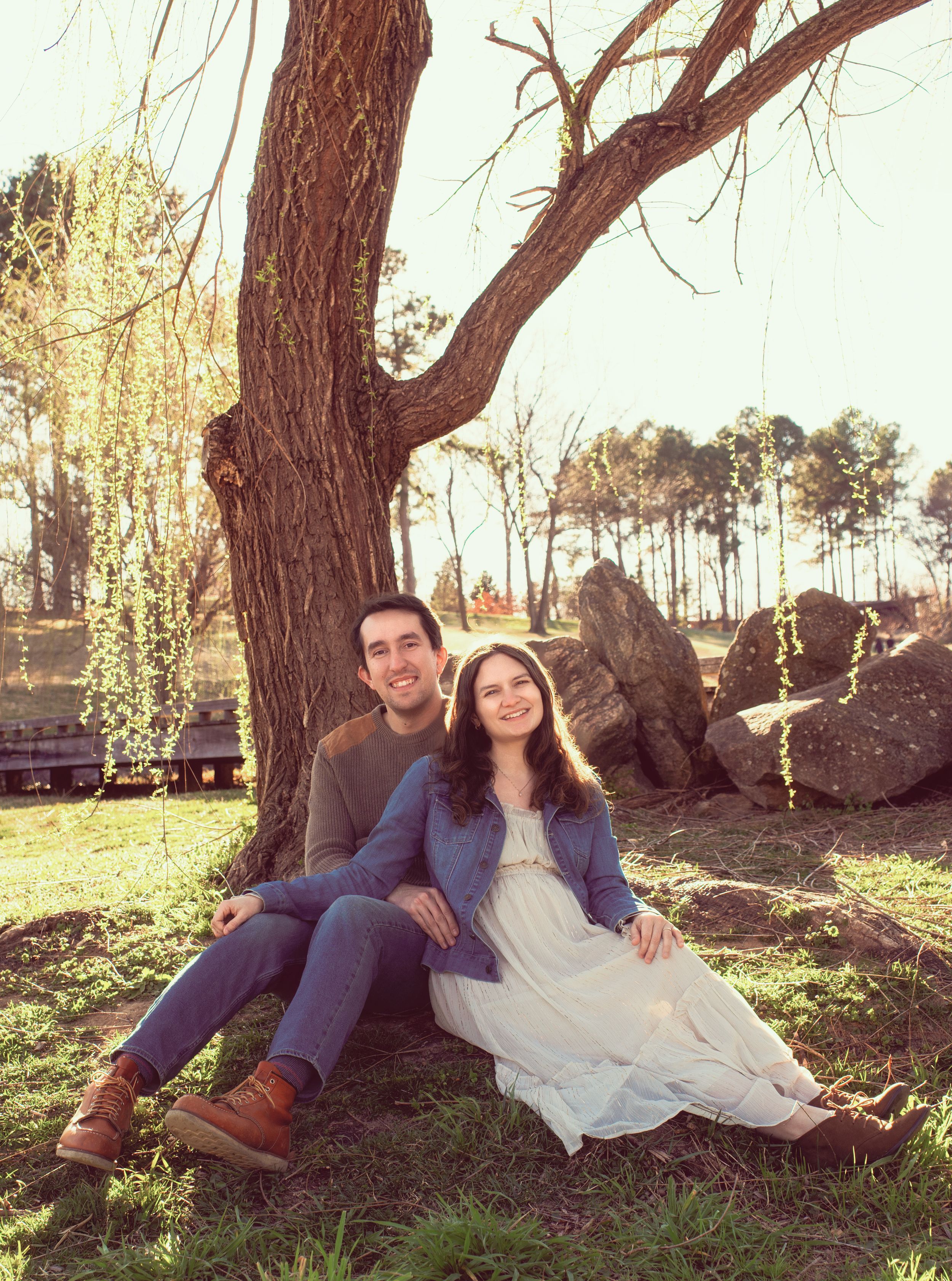 A couple relaxes together against a large tree trunk in a sunlit outdoor setting with a white dress and casual attire.