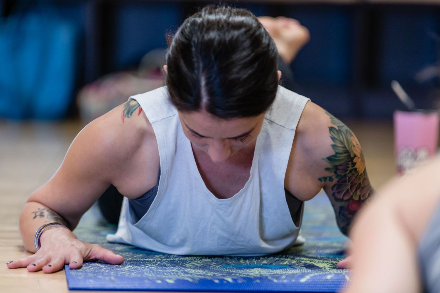 A person in a white tank top with visible tattoos leans over a yoga mat during practice at a studio.