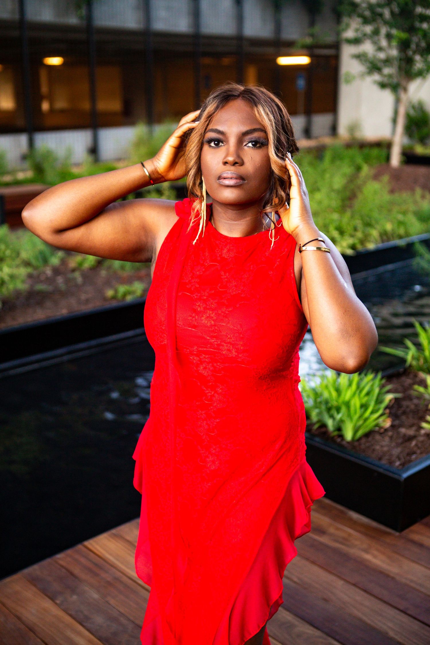 A woman in a vibrant red dress poses outdoors near landscaped planters at sunset.
