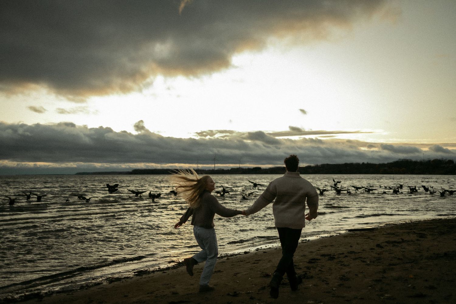Two silhouetted figures run along a beach shoreline at sunset with dramatic cloudy skies above.
