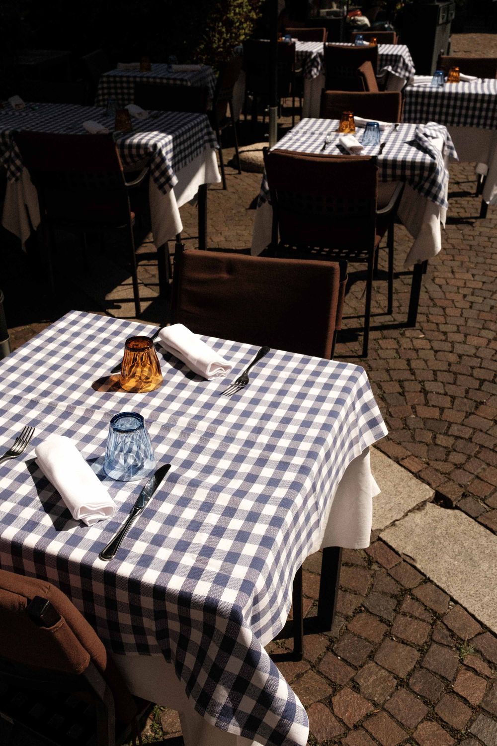 Outdoor cafe tables with blue and white checkered tablecloths set up on a brick patio in sunlight.