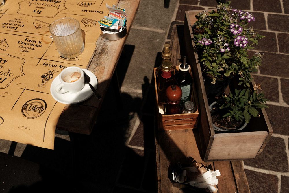 Sunlit outdoor table with teapot, drinks and potted flowers on brick pavement.