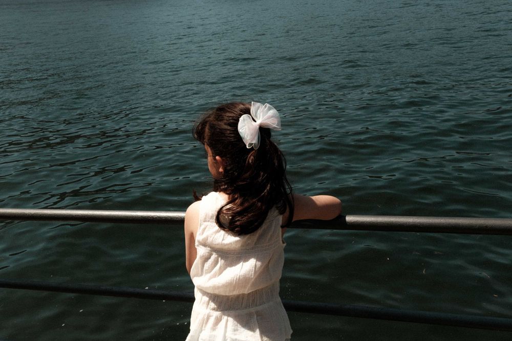 A figure in white clothing leans against a railing overlooking dark waters.