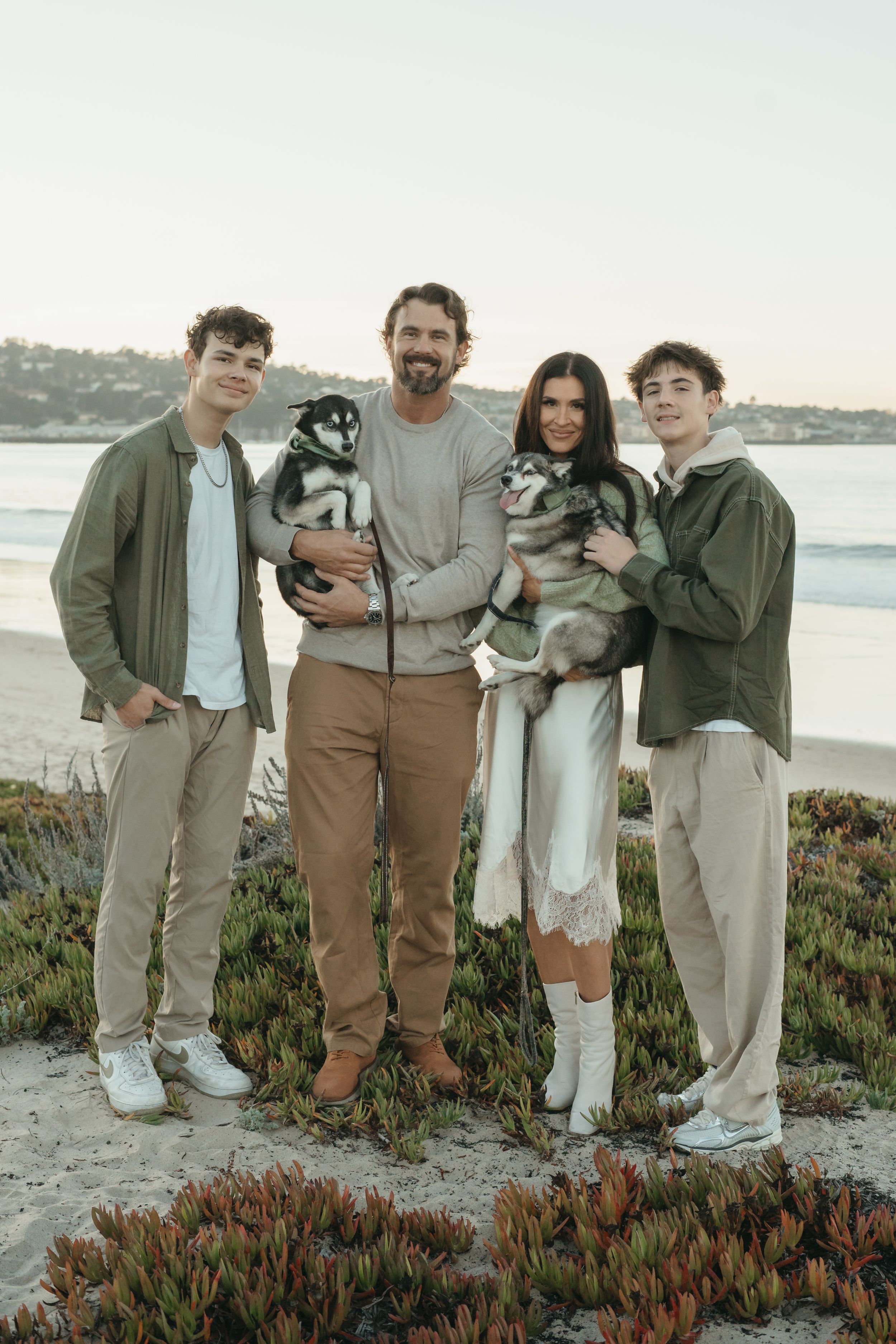 A family poses together with their dogs on a coastal bluff with green succulents and ocean views at sunset.