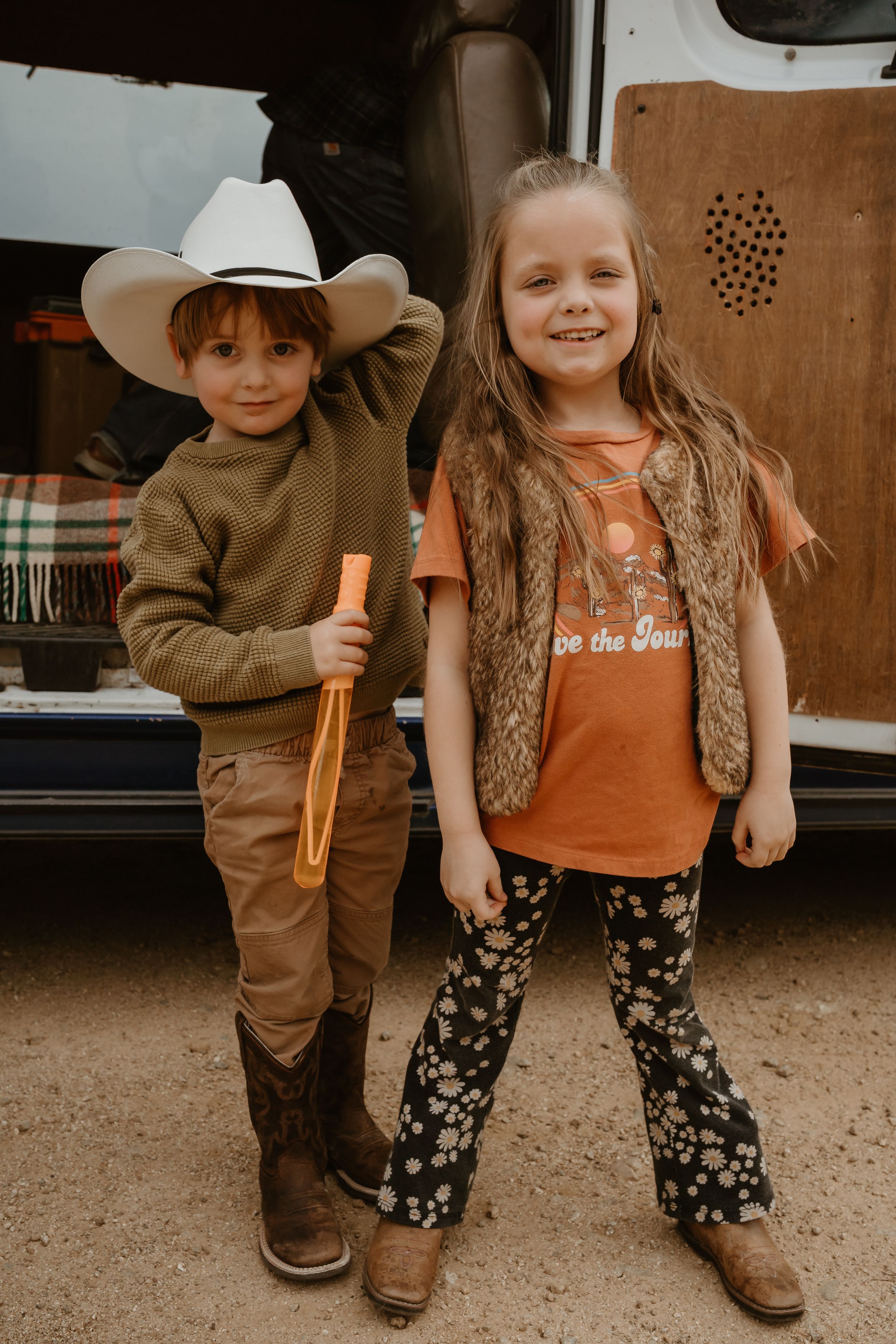 Two young children in western wear and cowboy boots pose together in front of a camper.
