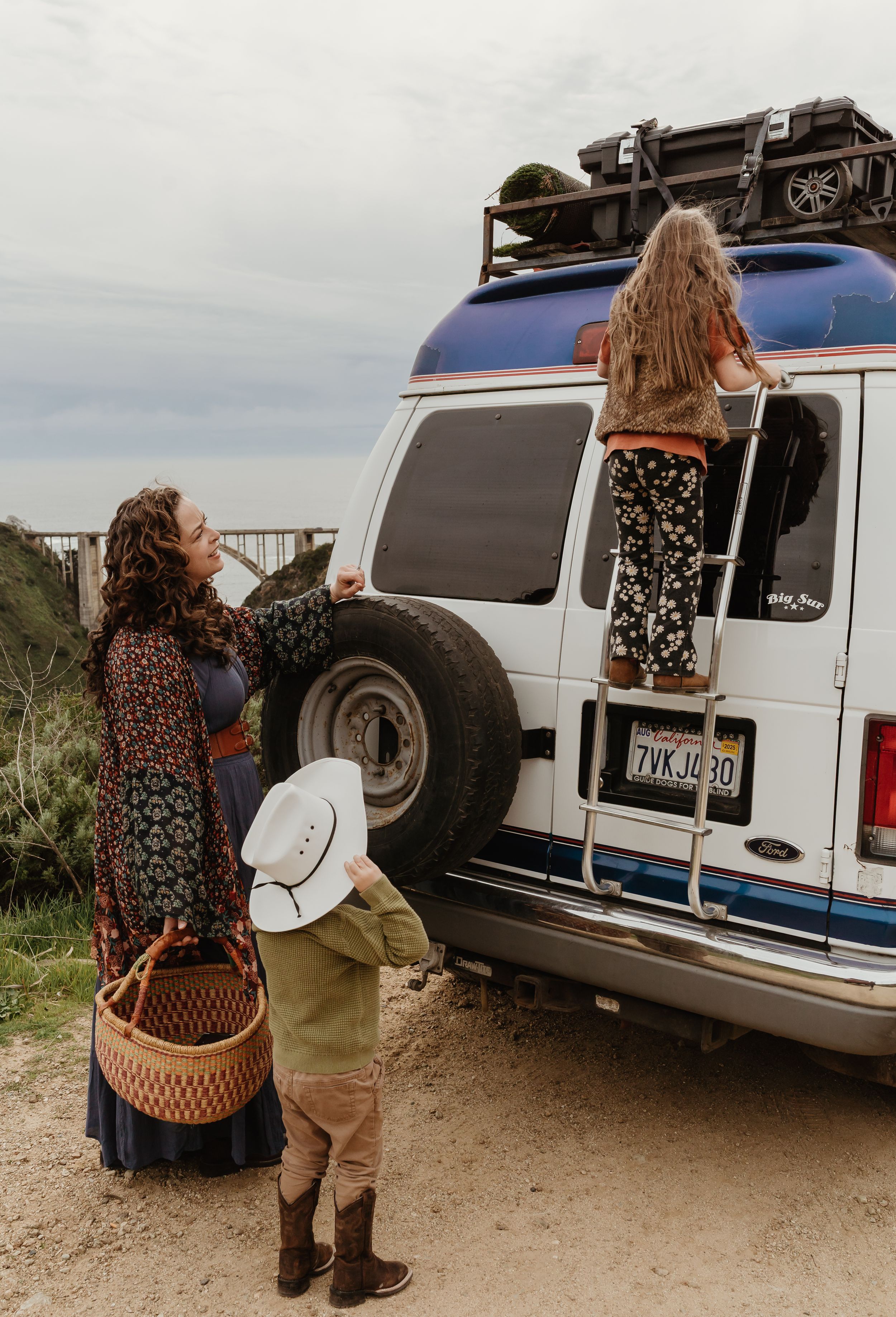 Adventure seekers in floral dresses gather around a white camper van with roof rack storage at a beach overlook.