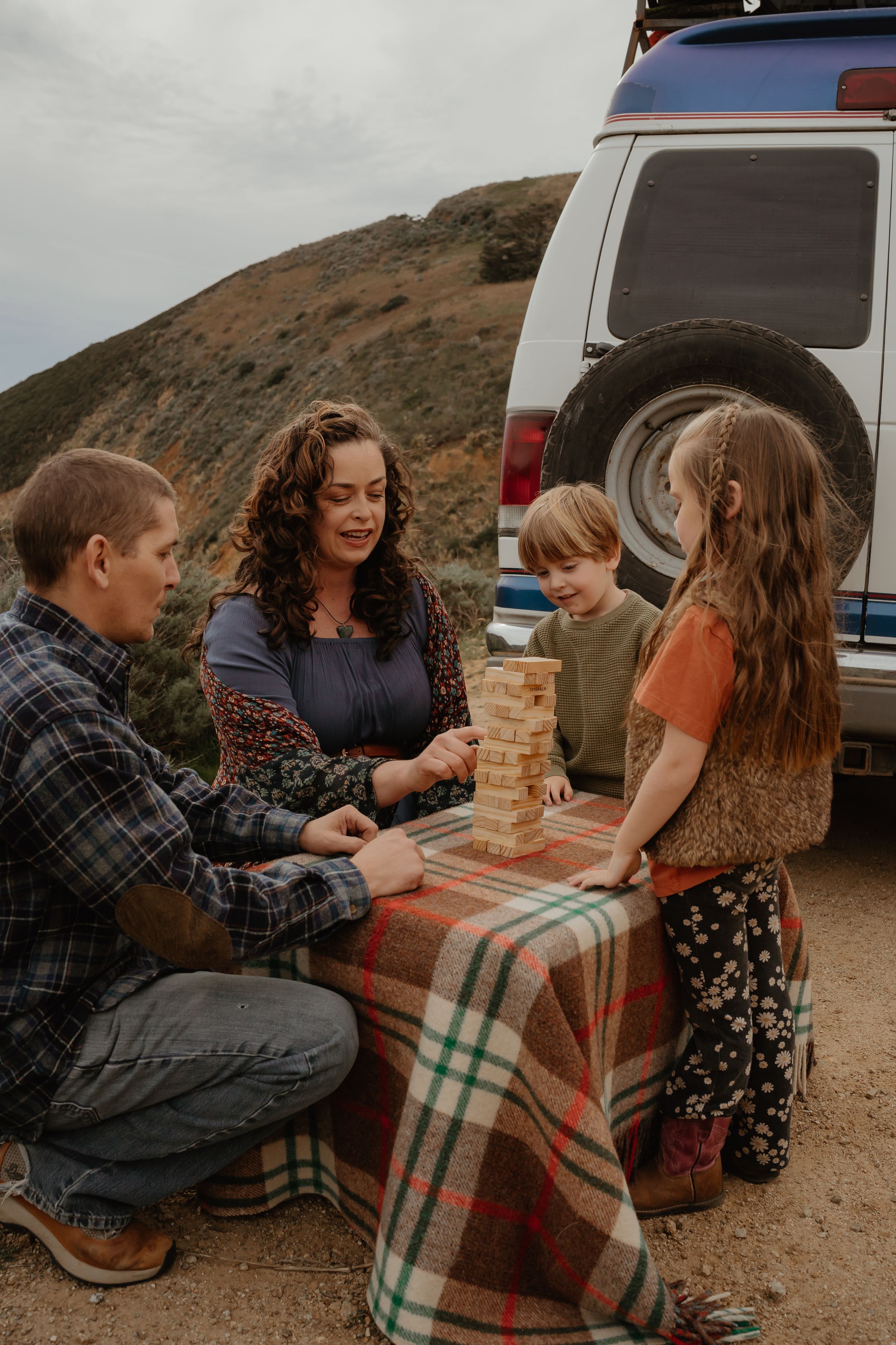 A family plays Jenga together on a plaid blanket while camping next to their SUV in the mountains.