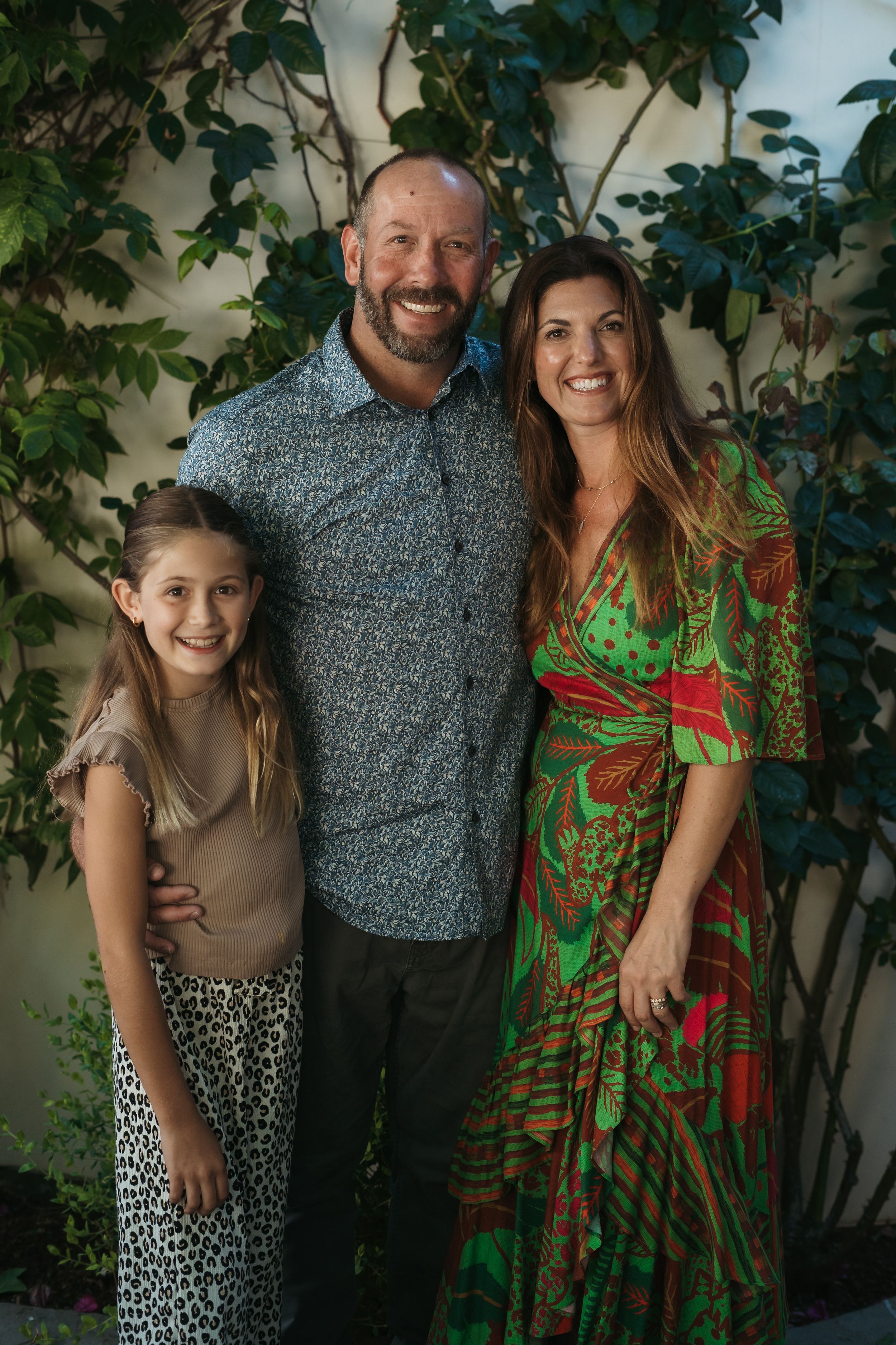 Happy family with daughter pose together against white wall with green climbing vines outdoors.